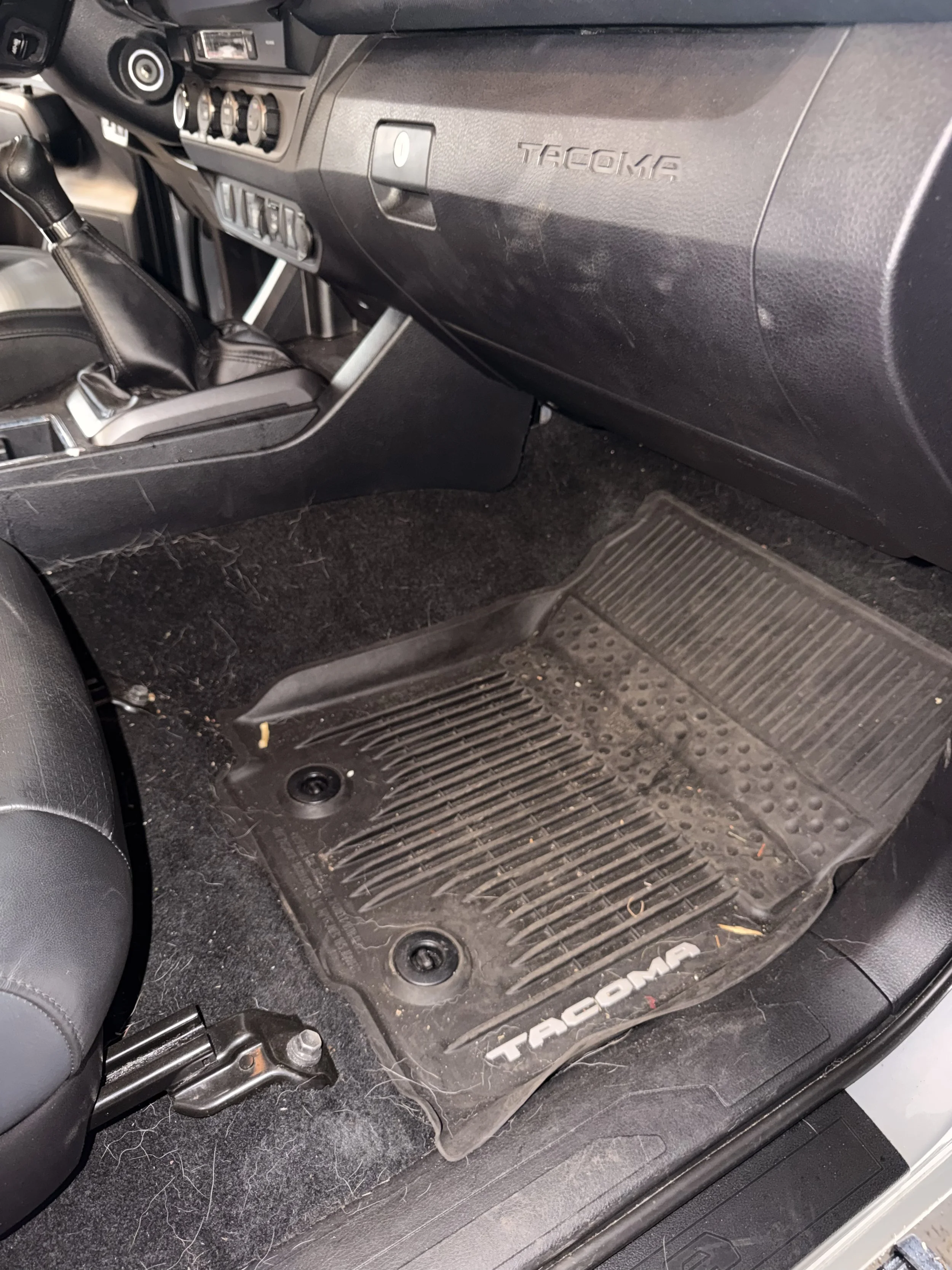 Interior view of a vehicle showing the driver's side floor mat, which is dirty with hair, debris, and some small objects. The dashboard and gear shift are partially visible, and there is a black rubber floor mat with the 