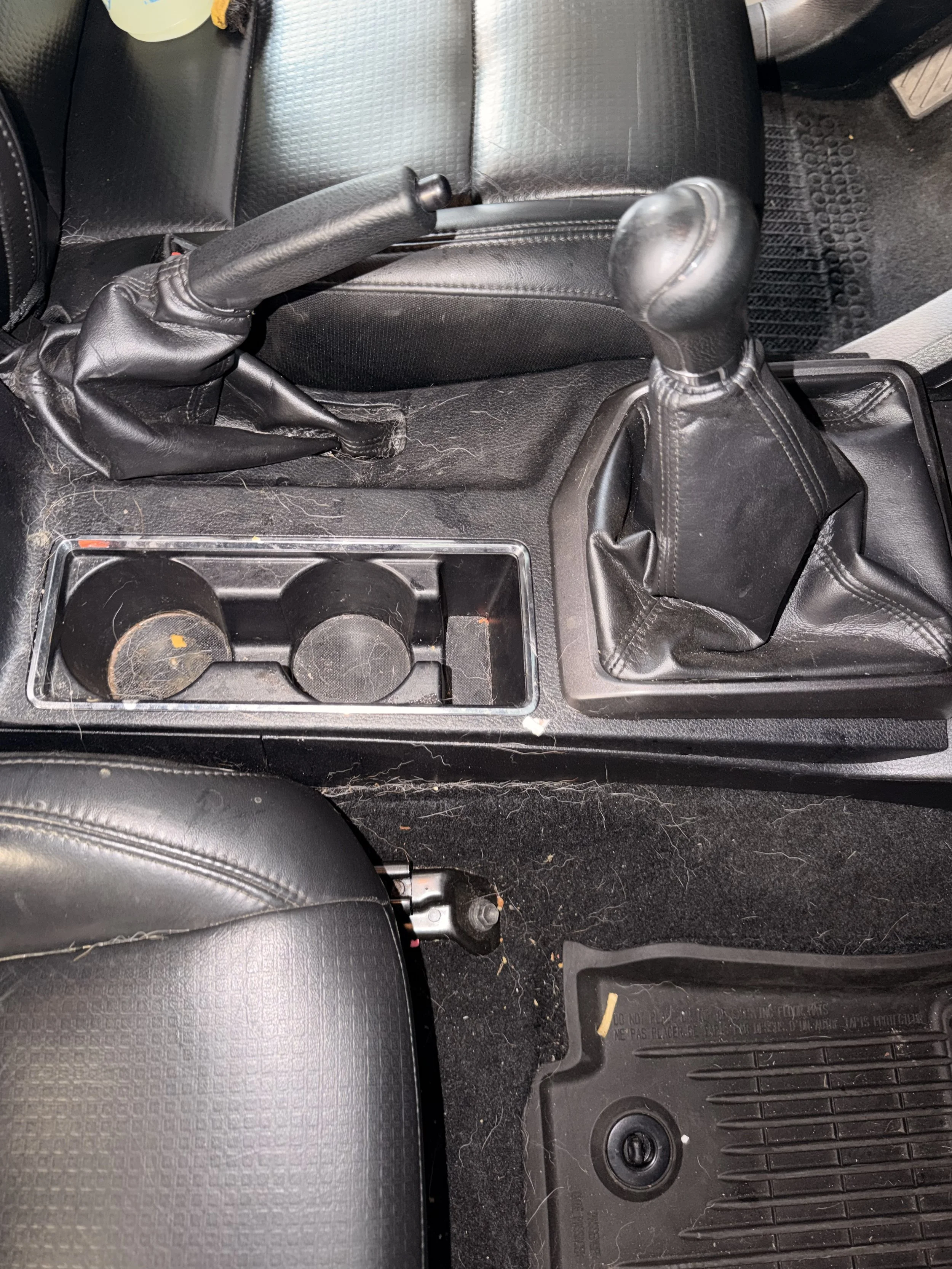 The interior of a vehicle showing a gear shift, handbrake, and cup holder area with some dirt and debris on the floor.
