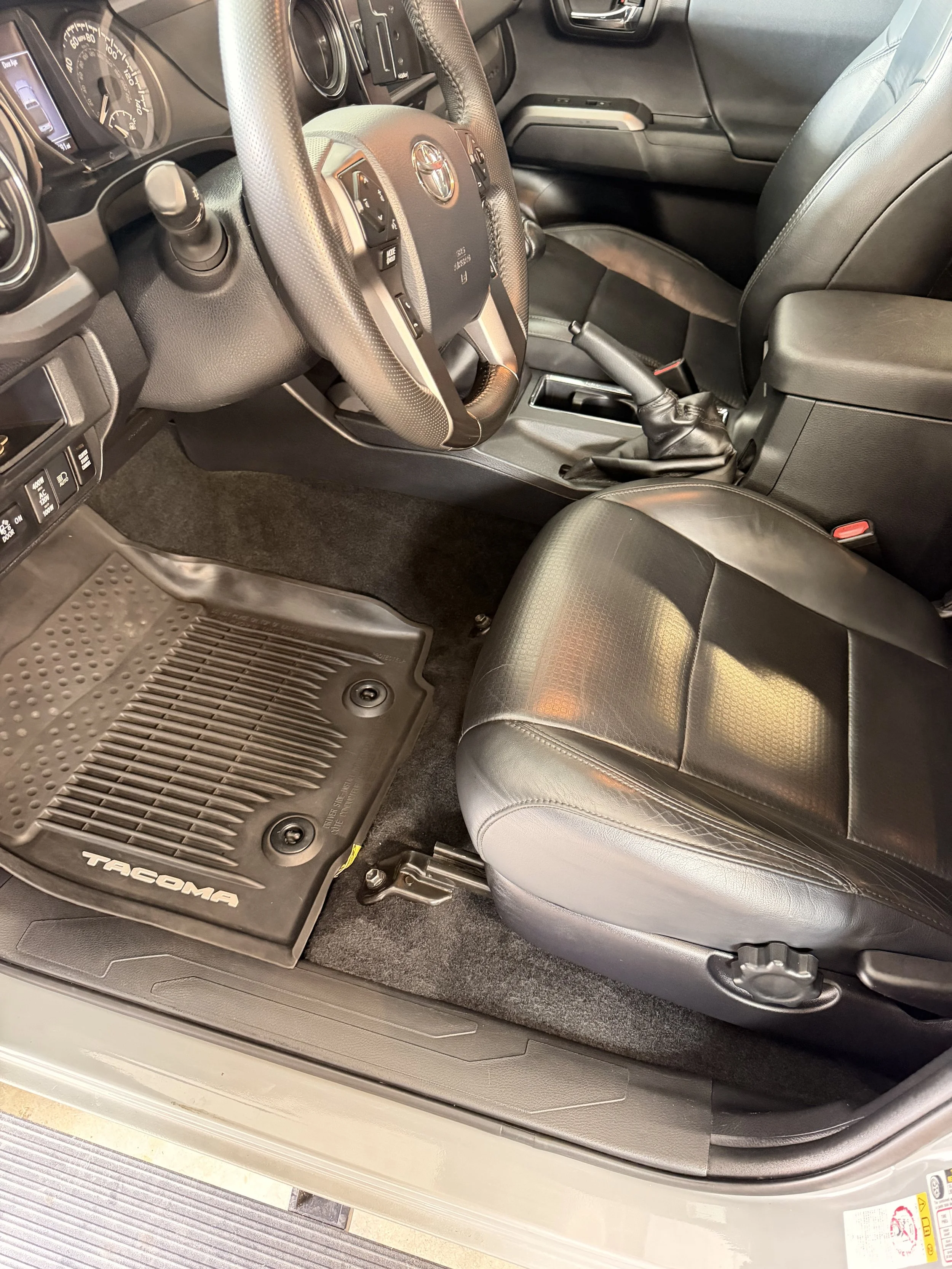 Interior of a Toyota Tacoma truck showing the driver's seat, steering wheel, dashboard, gear shift, and floor mat.