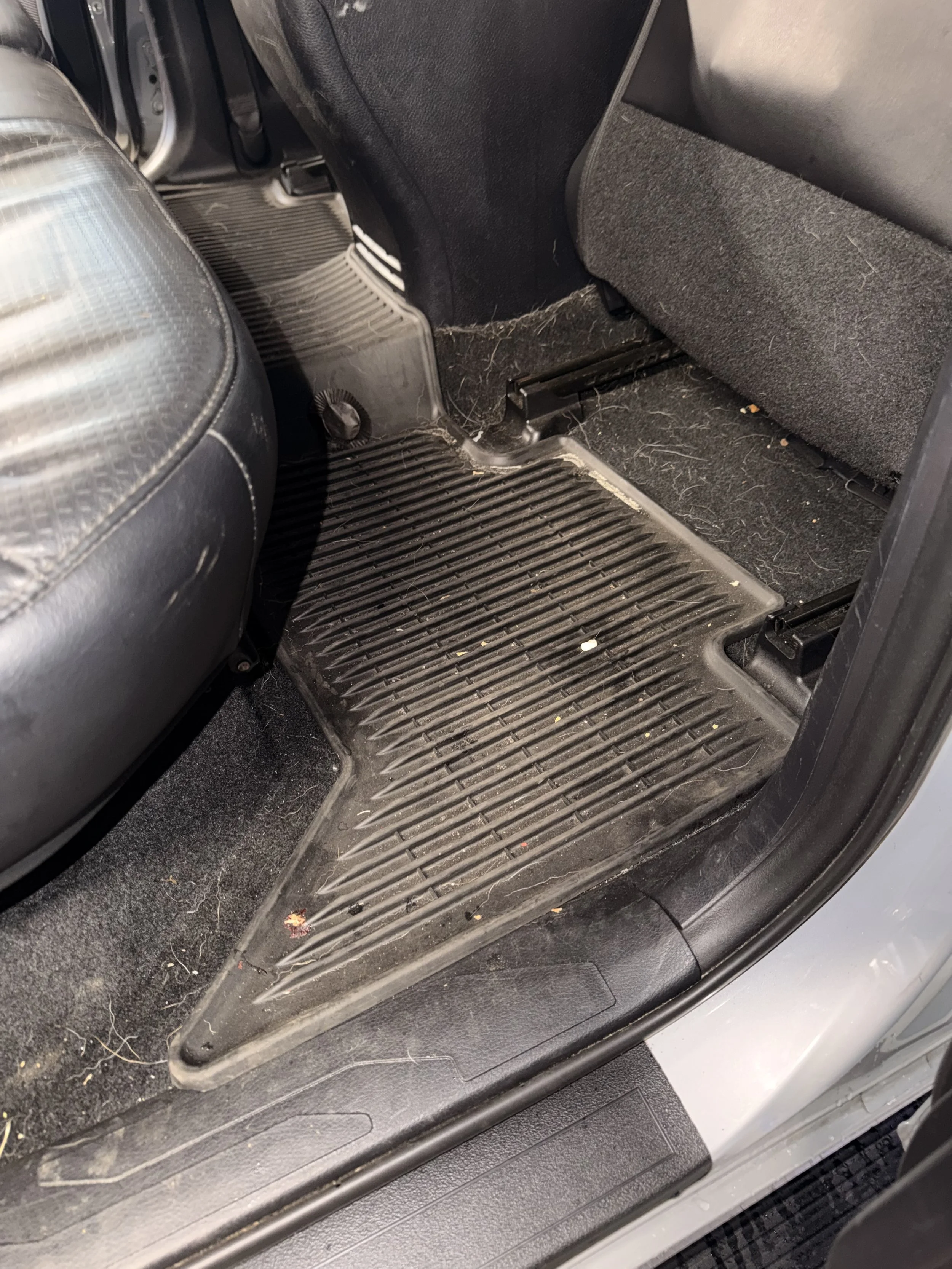 The interior of a car showing the footwell area with a black rubber floor mat, some dirt and debris, and part of a black seat and door panel.
