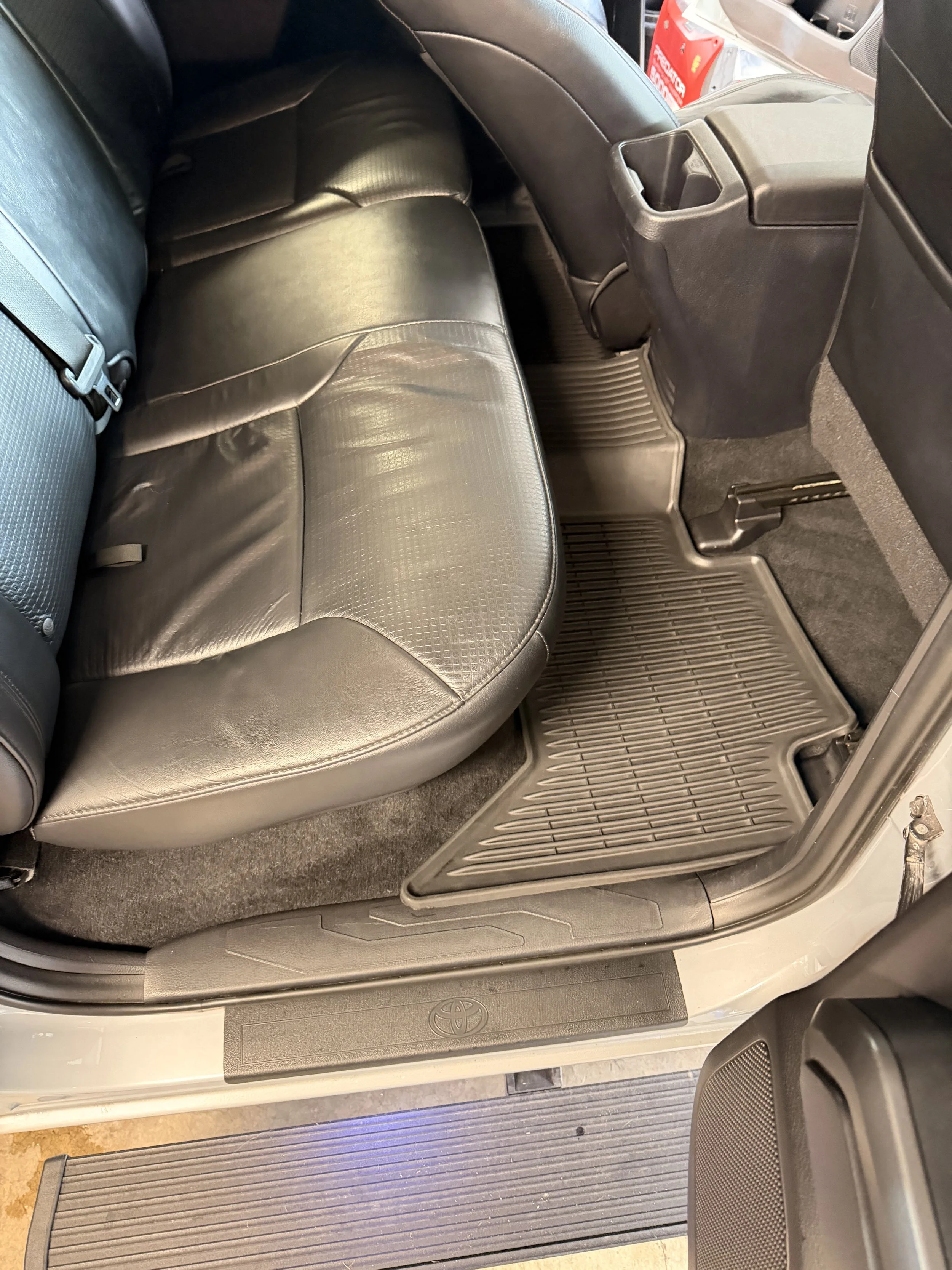 The interior of a vehicle showing a black leather backseat, black floor mats, and gray side panels. The front passenger side door is open, revealing a gray floor mat and part of the door panel.