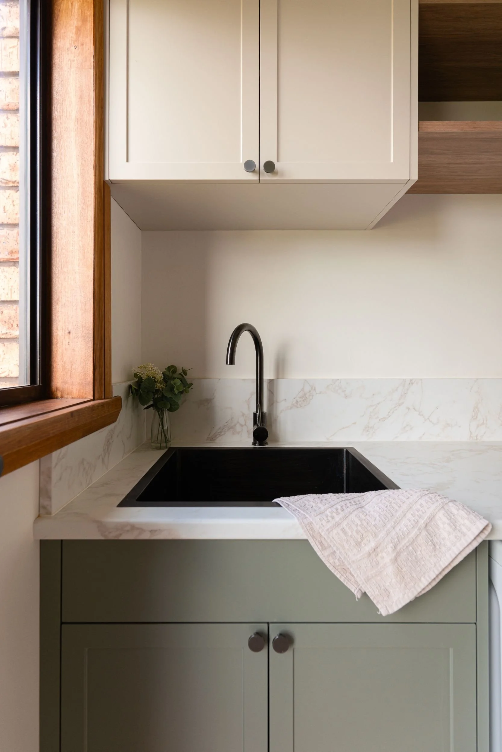 Laundry sink with gunmetal tapware and soft green cabinetry beside a timber window
