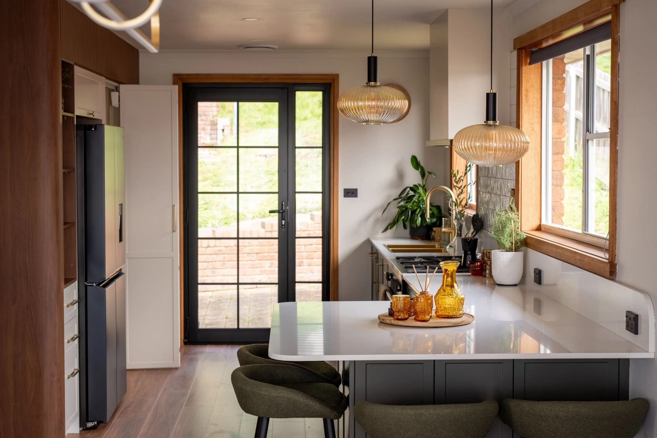 Mid-century inspired kitchen with dark cabinetry, white stone island, amber glass pendants and black-framed French doors, Riverside Tasmania