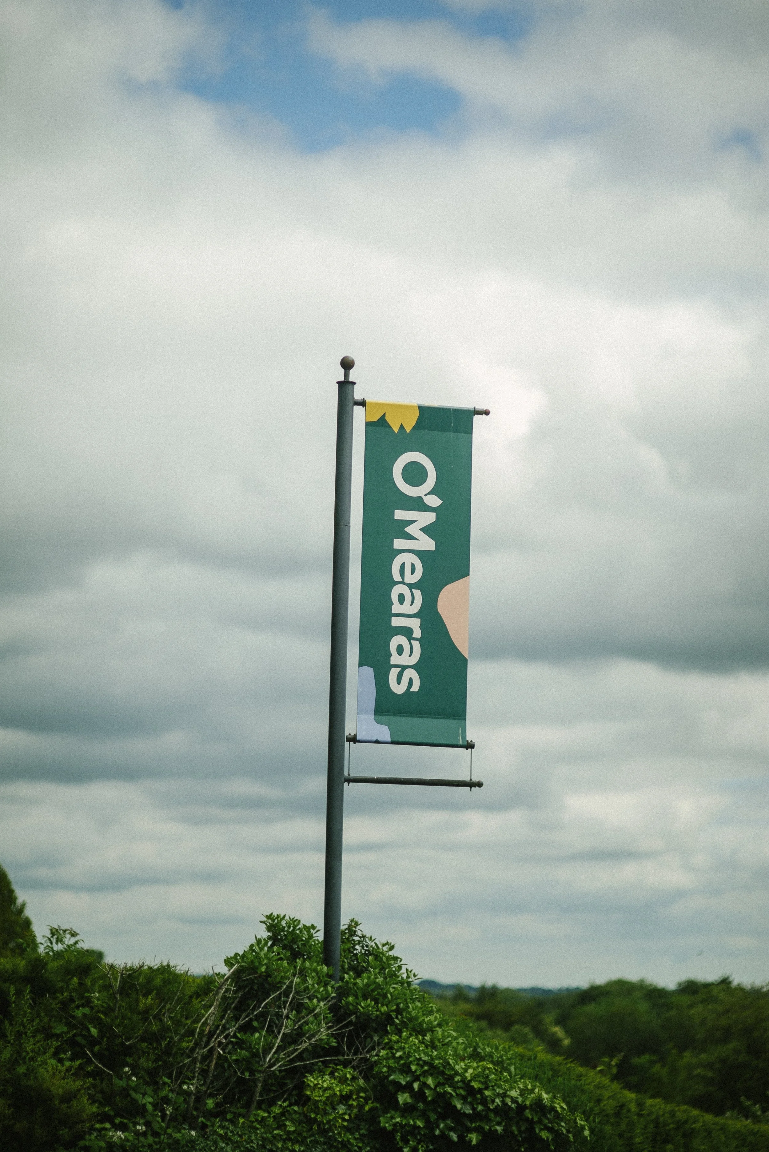 A Q Meadows flag banner hanging from a tall pole outdoors with green bushes and a cloudy sky in the background.