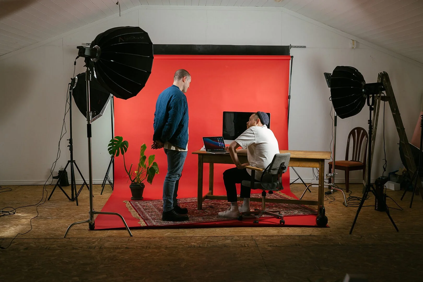 Two men in a photography studio with a red backdrop, one standing and one sitting at a desk with a laptop. There are studio lights and a potted plant on a patterned rug.