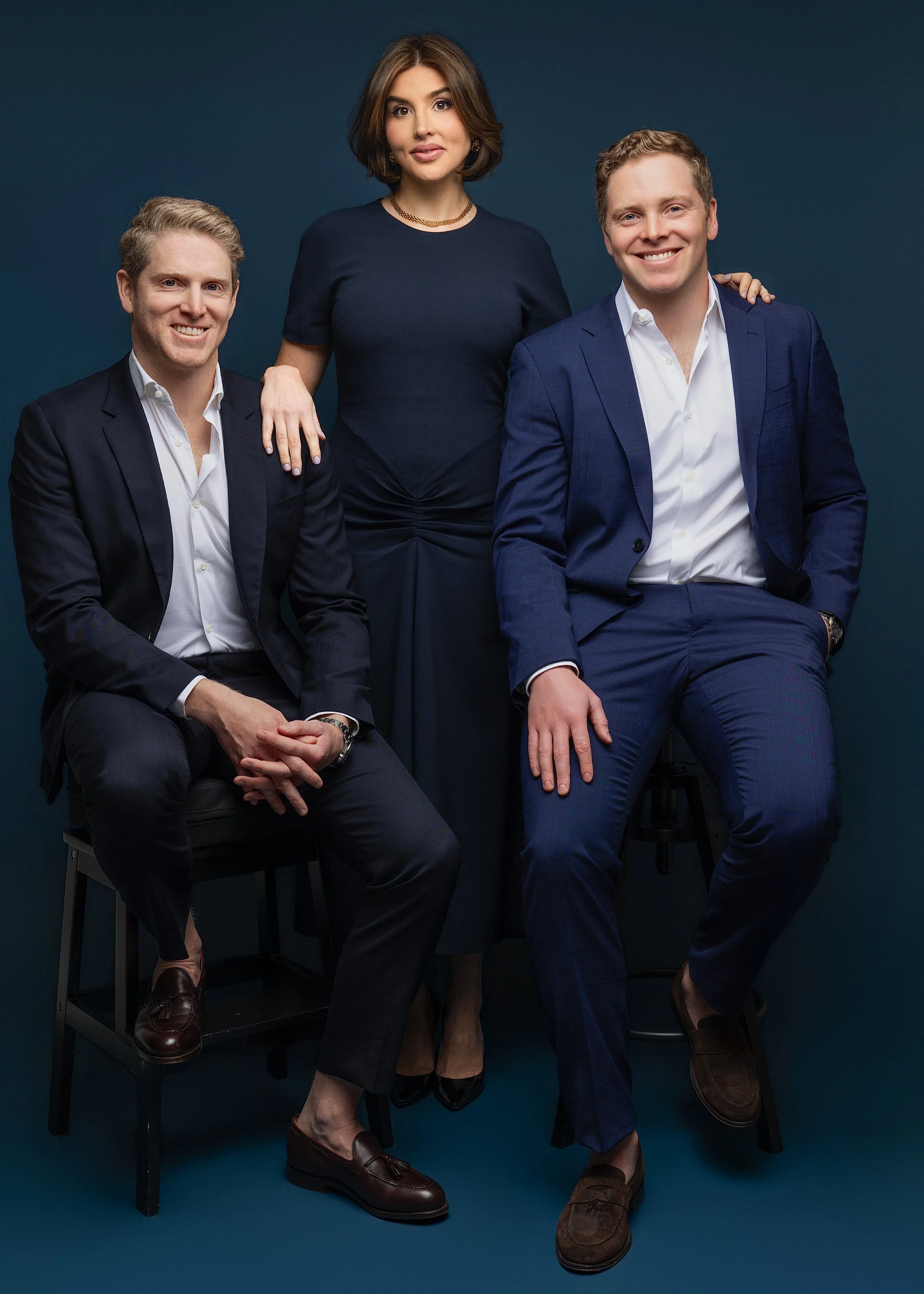 Three well-dressed individuals, two men seated and one woman standing behind them, posing against a dark blue background.
