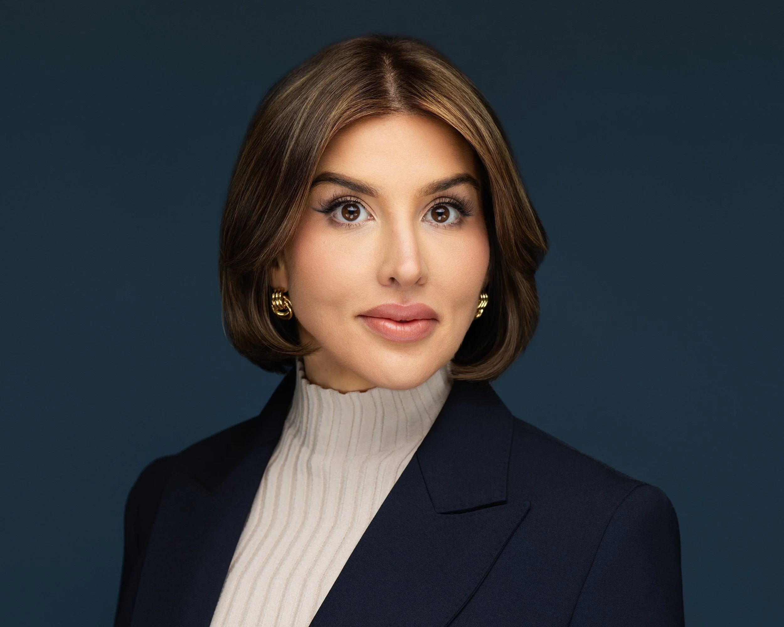 A professional woman with shoulder-length brown hair, wearing a beige turtleneck sweater and a dark blazer, with gold earrings, against a dark blue background.