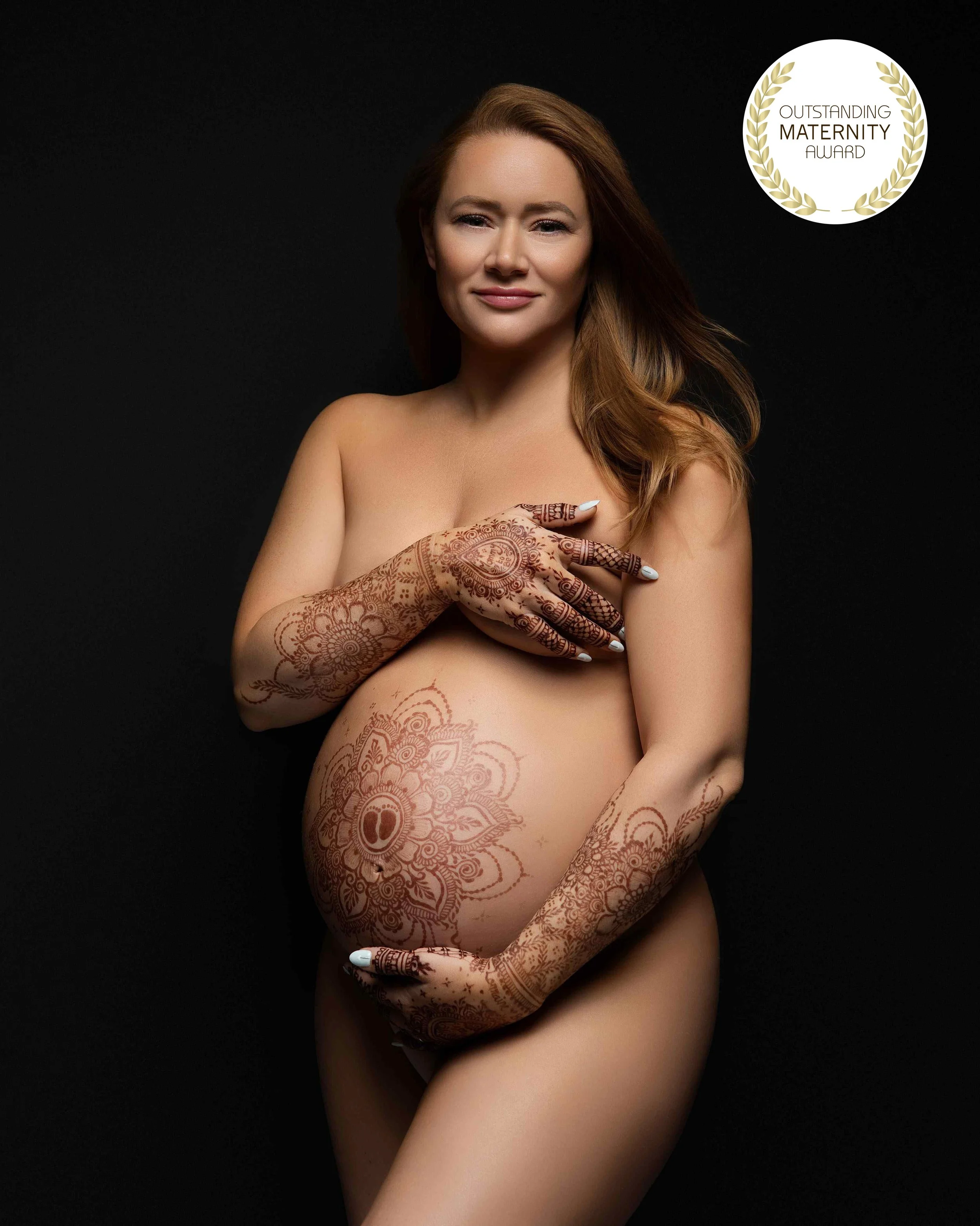 Nude pregnant woman with mehndi on arms, shoulders, and abdomen, smiling with right hand over her chest, black background, 'Outstanding Maternity Award' badge in upper right corner.