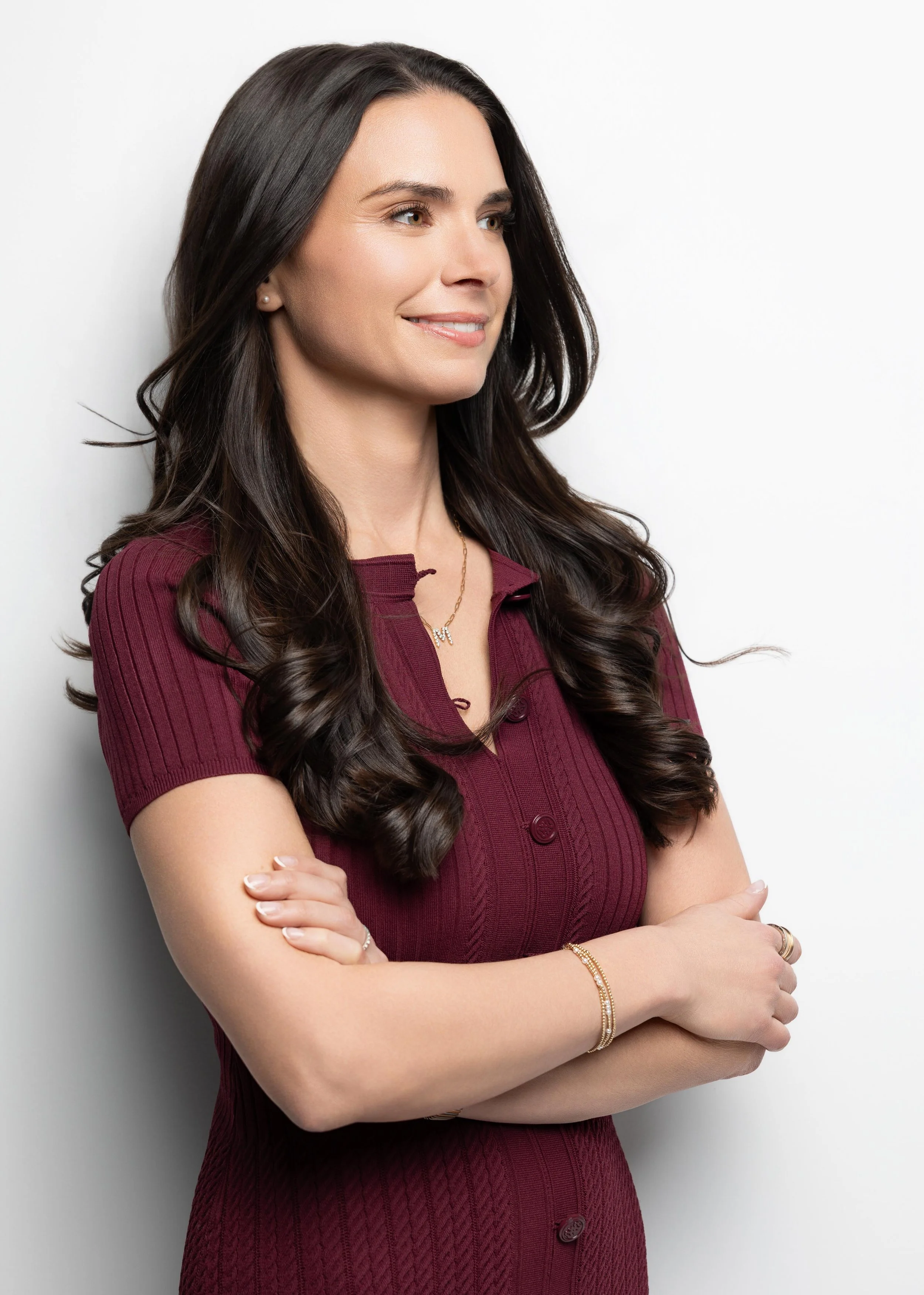 A woman with long, dark hair styled in loose waves is standing with crossed arms against a plain white background. She is wearing a short-sleeved, maroon, ribbed knit dress, gold jewelry including a necklace and bracelets, and has a slight smile on h