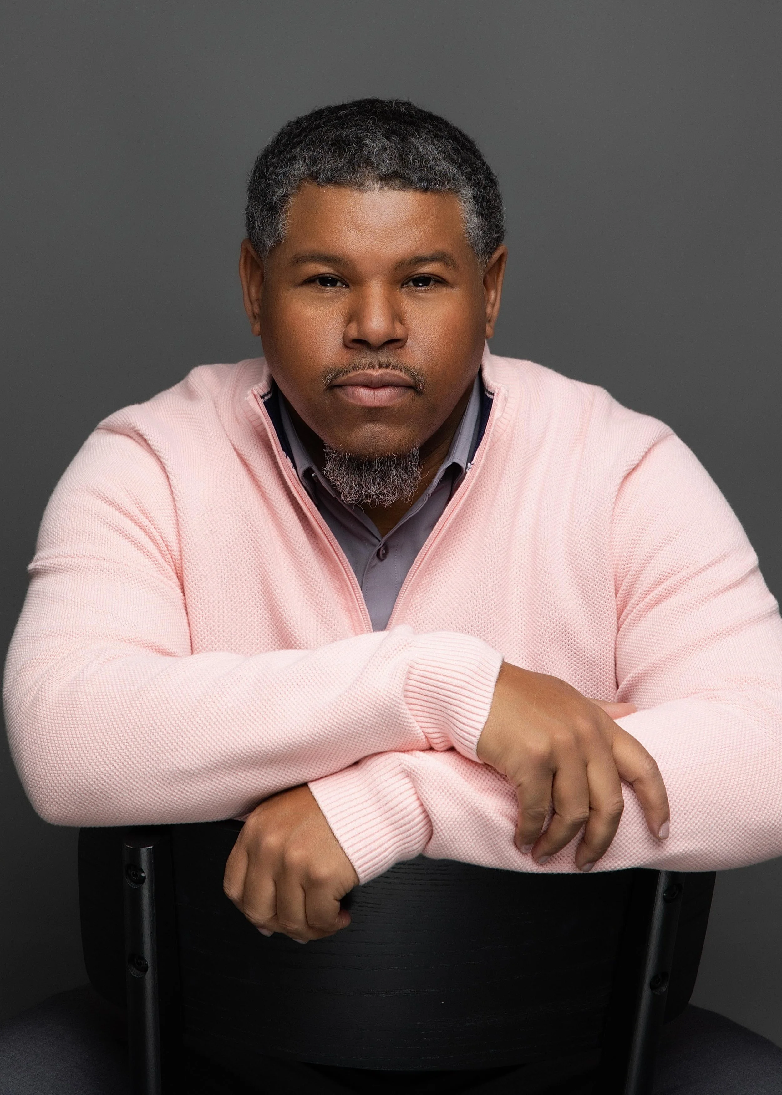 A man with short, curly gray hair and a gray goatee, wearing a pink sweater over a gray collared shirt, sitting with arms crossed on the back of a black chair against a dark gray background.