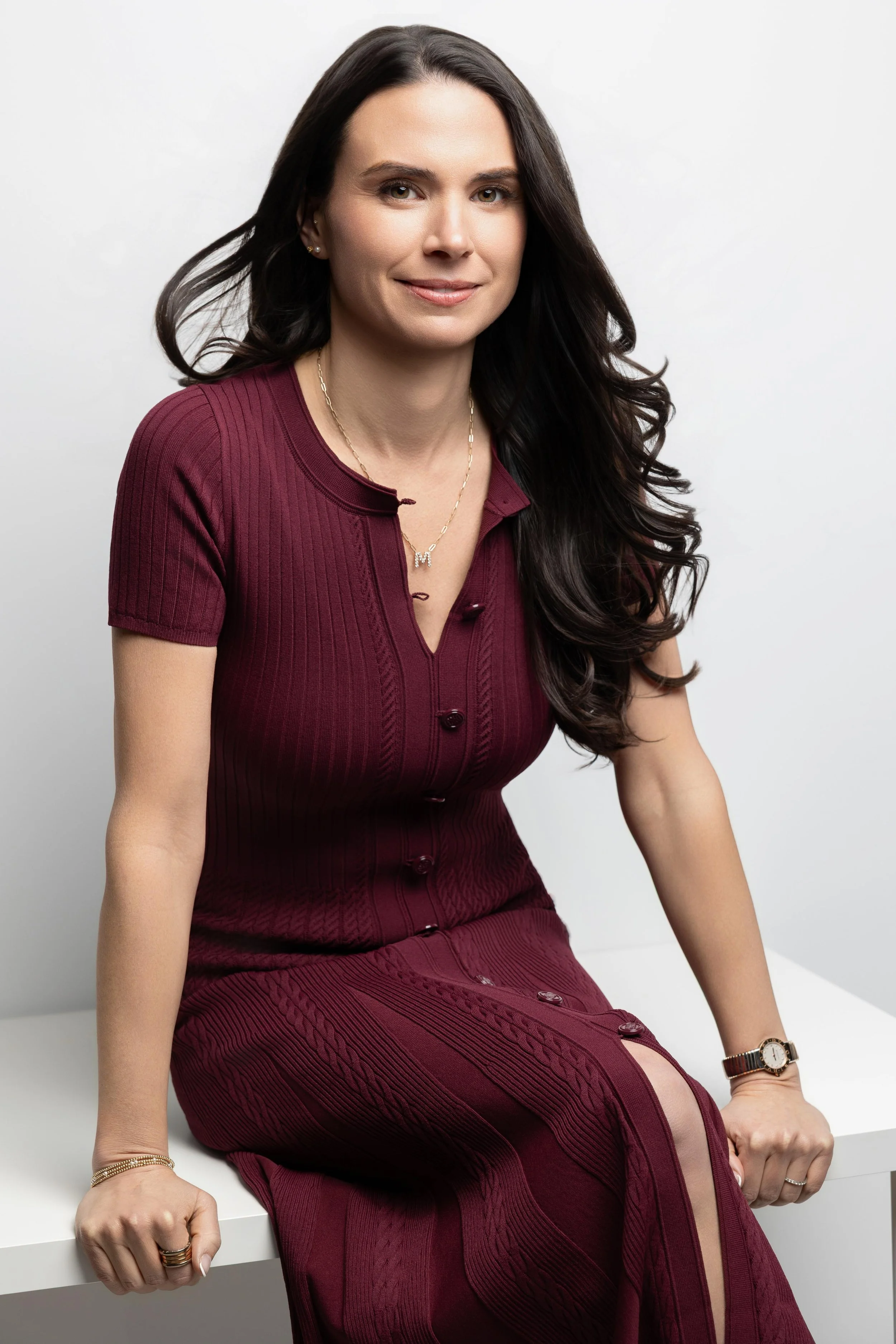 A woman with long dark hair wearing a maroon dress and a watch is sitting on a white surface against a plain white background.