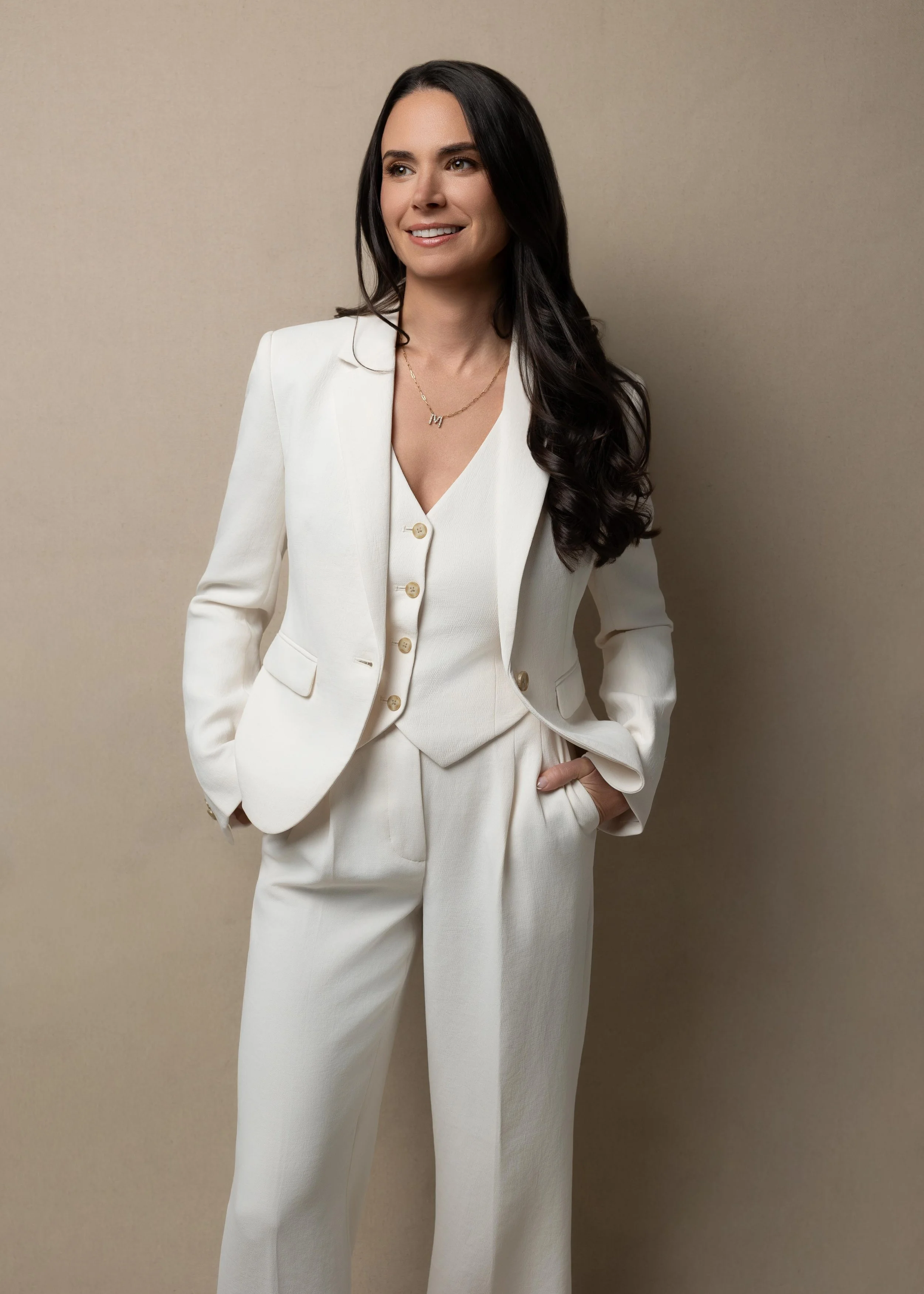 A woman with long dark hair wearing a cream-colored pantsuit and smiling, standing against a plain beige background.