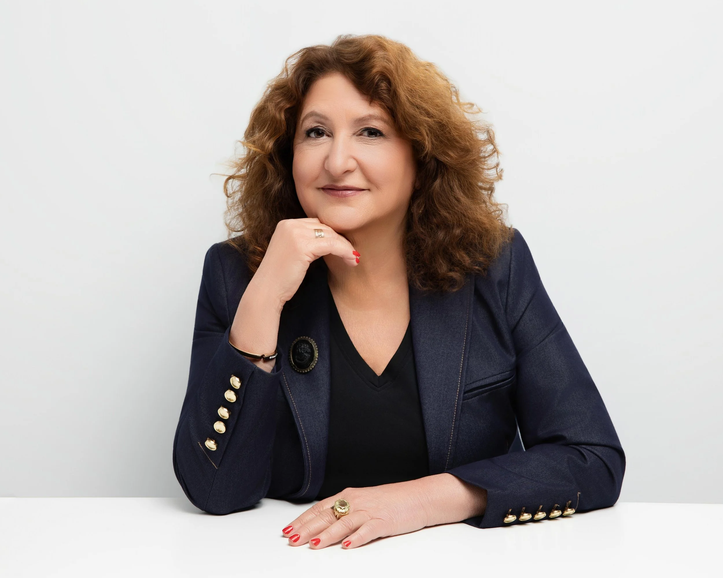 middle-aged woman with curly red hair, wearing a navy blazer with gold buttons, sitting at a table with a white background, resting her chin on her right hand, smiling gently.