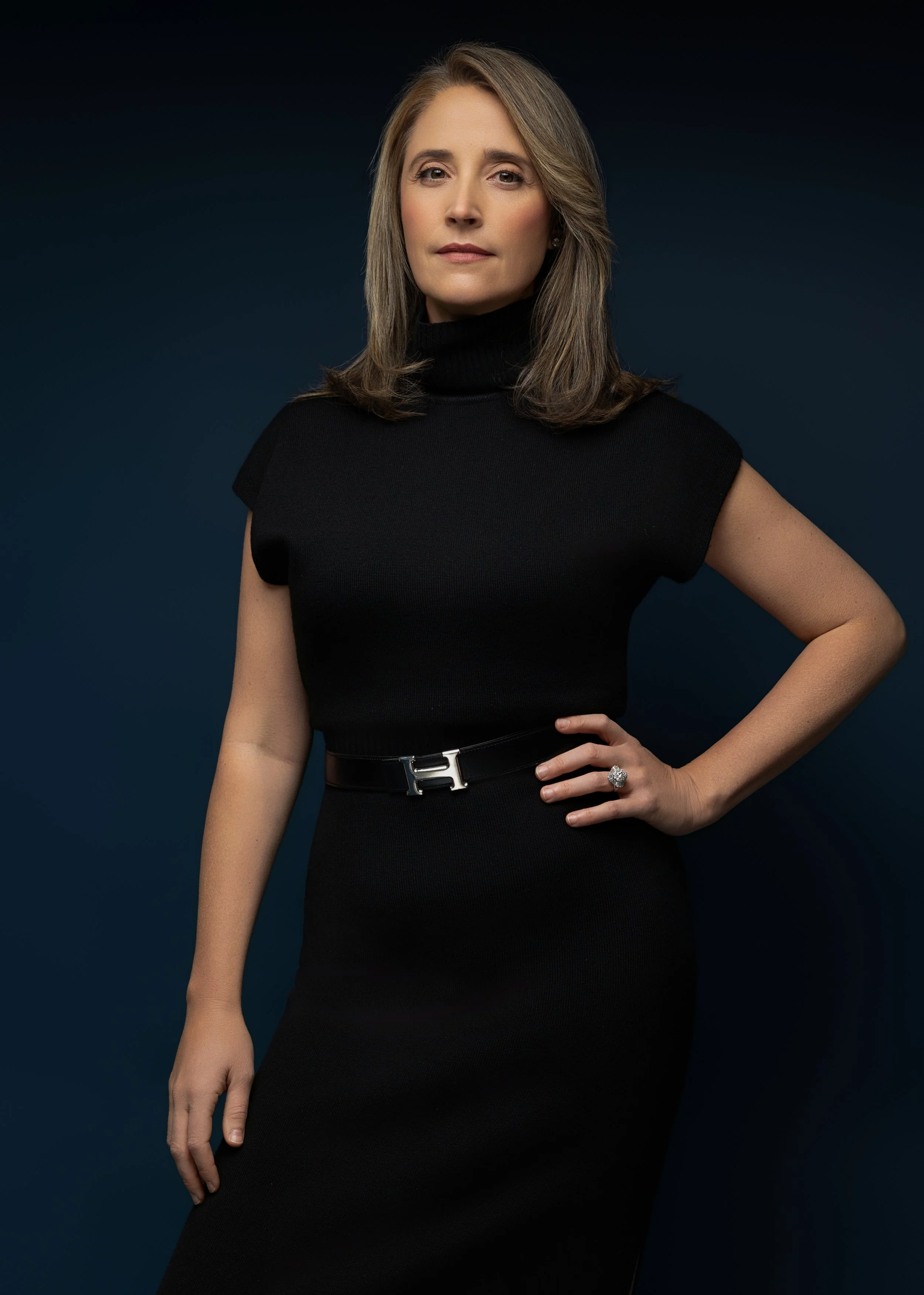 A woman with shoulder-length light brown hair, wearing a black turtleneck dress with short sleeves and a black belt, standing against a dark background.
