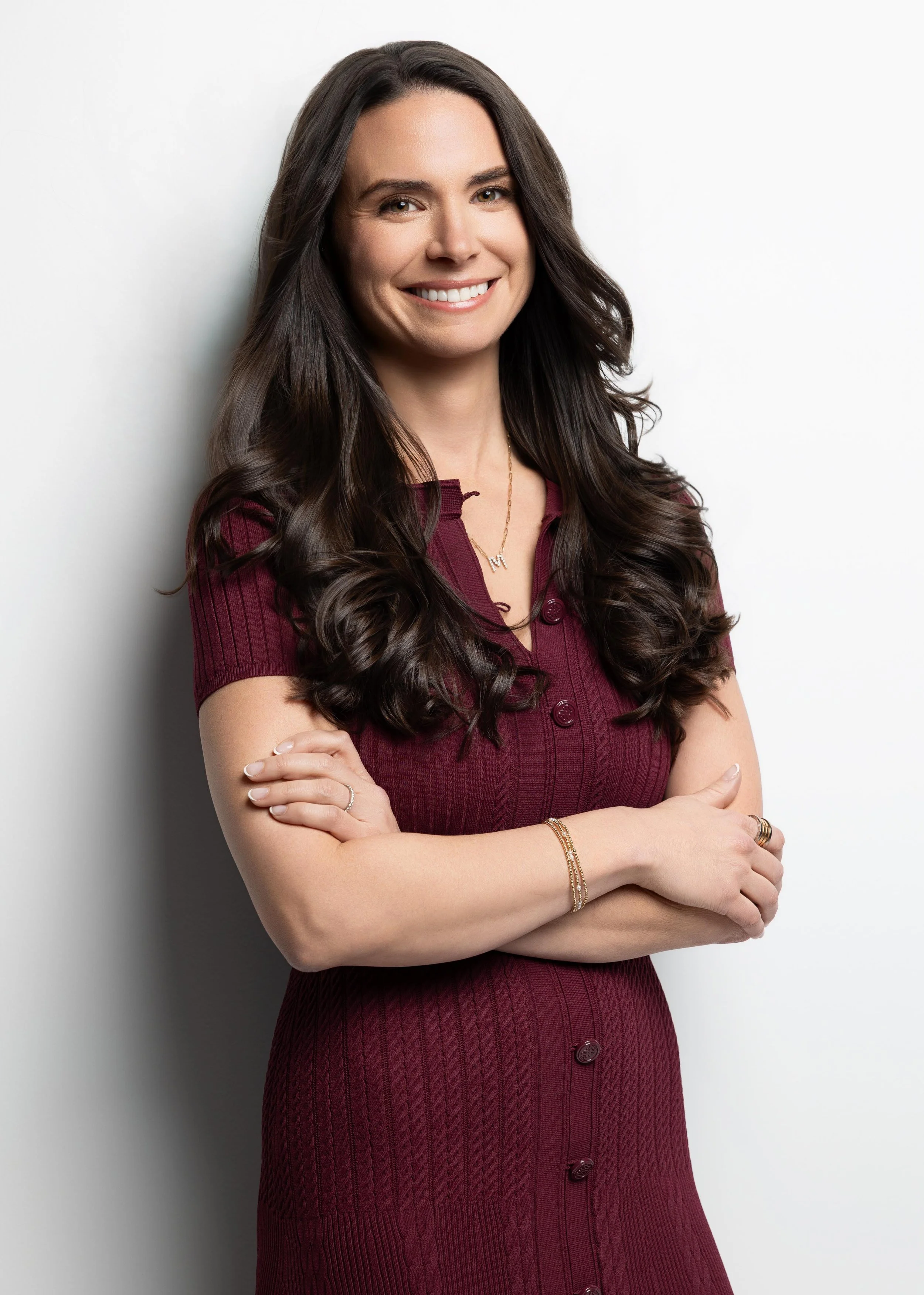 A woman with long dark brown hair, smiling, wearing a maroon button-up dress and gold jewelry, standing against a white background.
