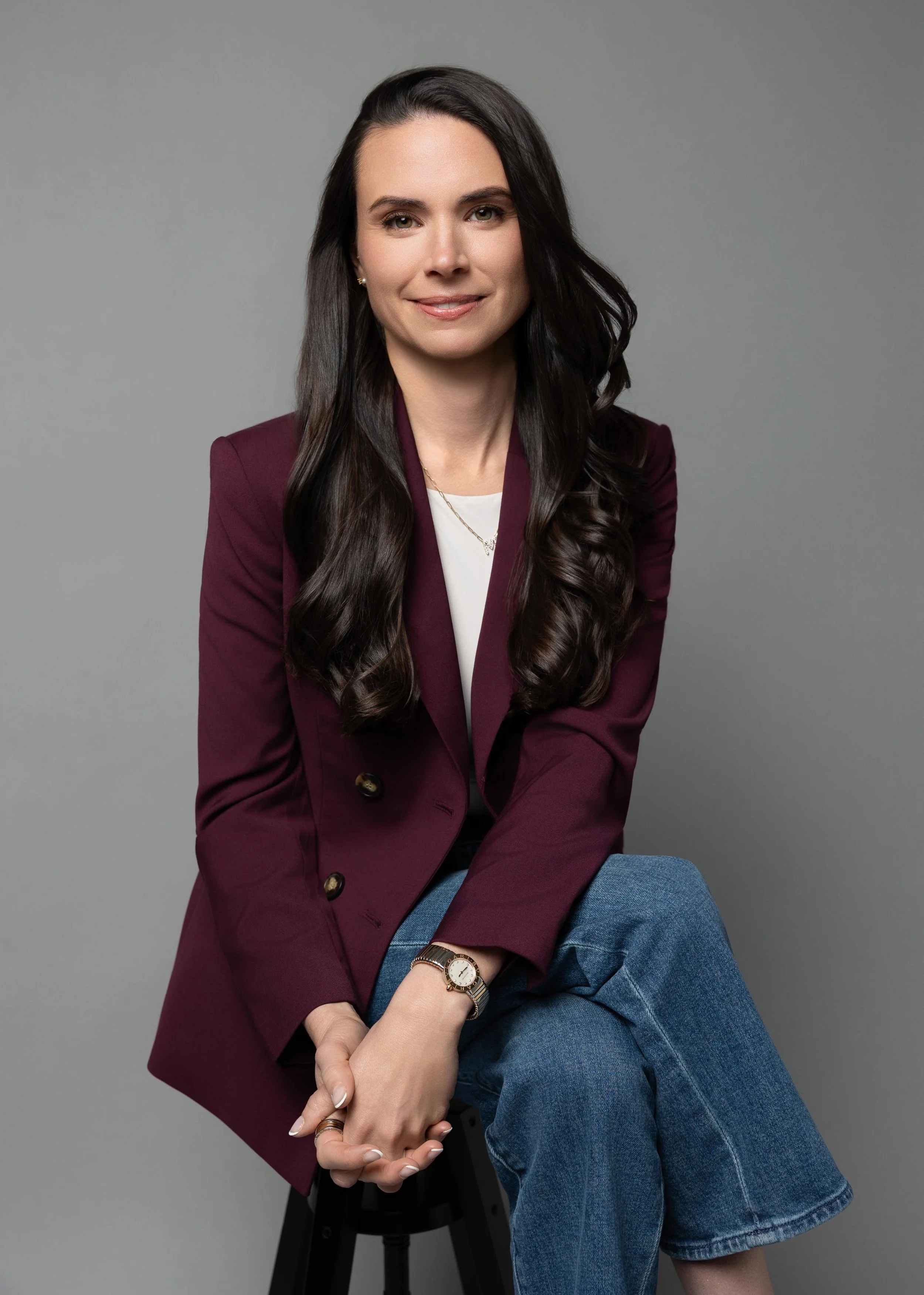 A woman with long, dark, wavy hair wearing a burgundy blazer, white top, and blue jeans, seated against a plain gray background.