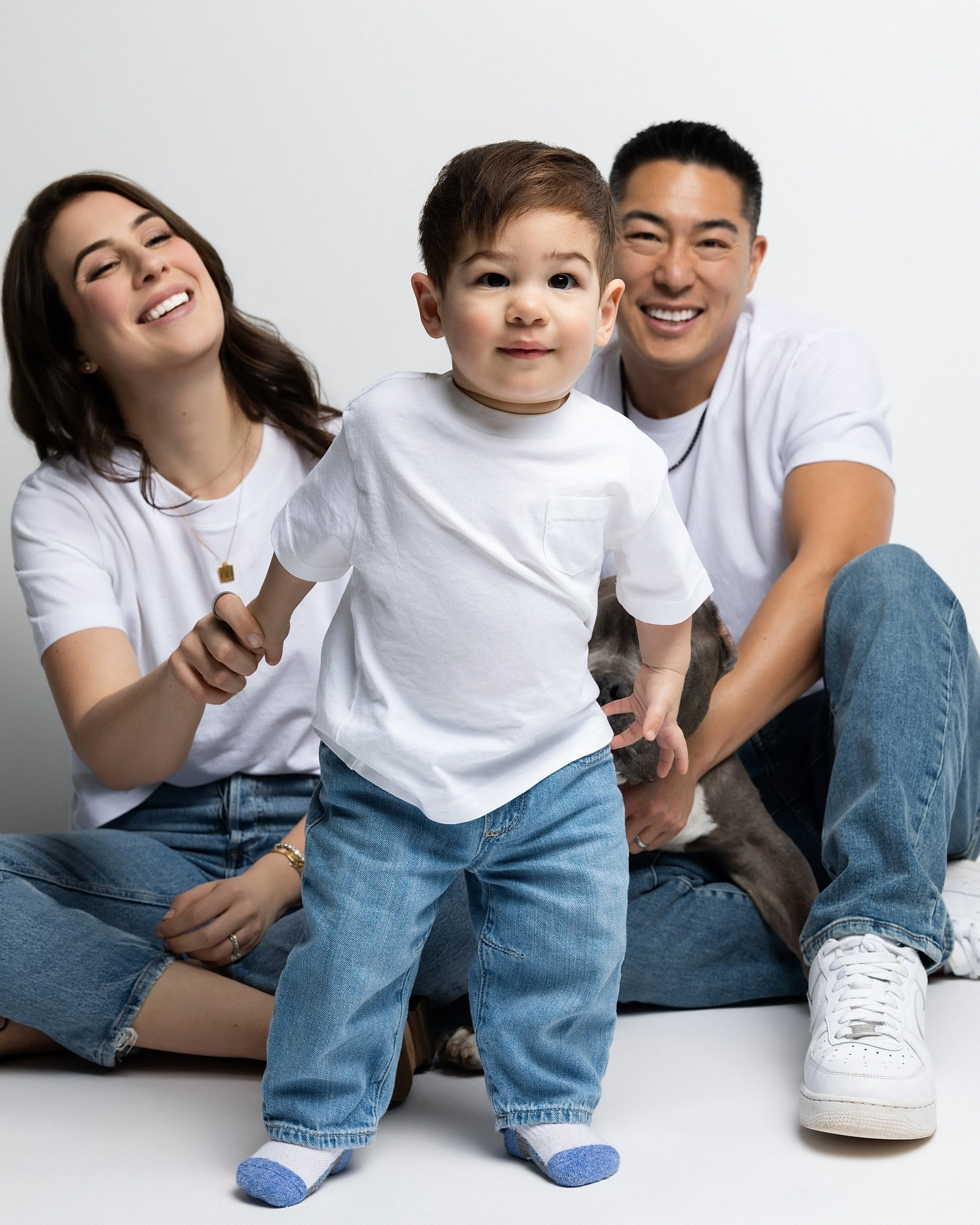 A happy family of three with their puppy, sitting on the floor against a white background. The woman has shoulder-length brown hair and is smiling. The man has short black hair and is smiling, wearing a white t-shirt and jeans. The young boy, dressed