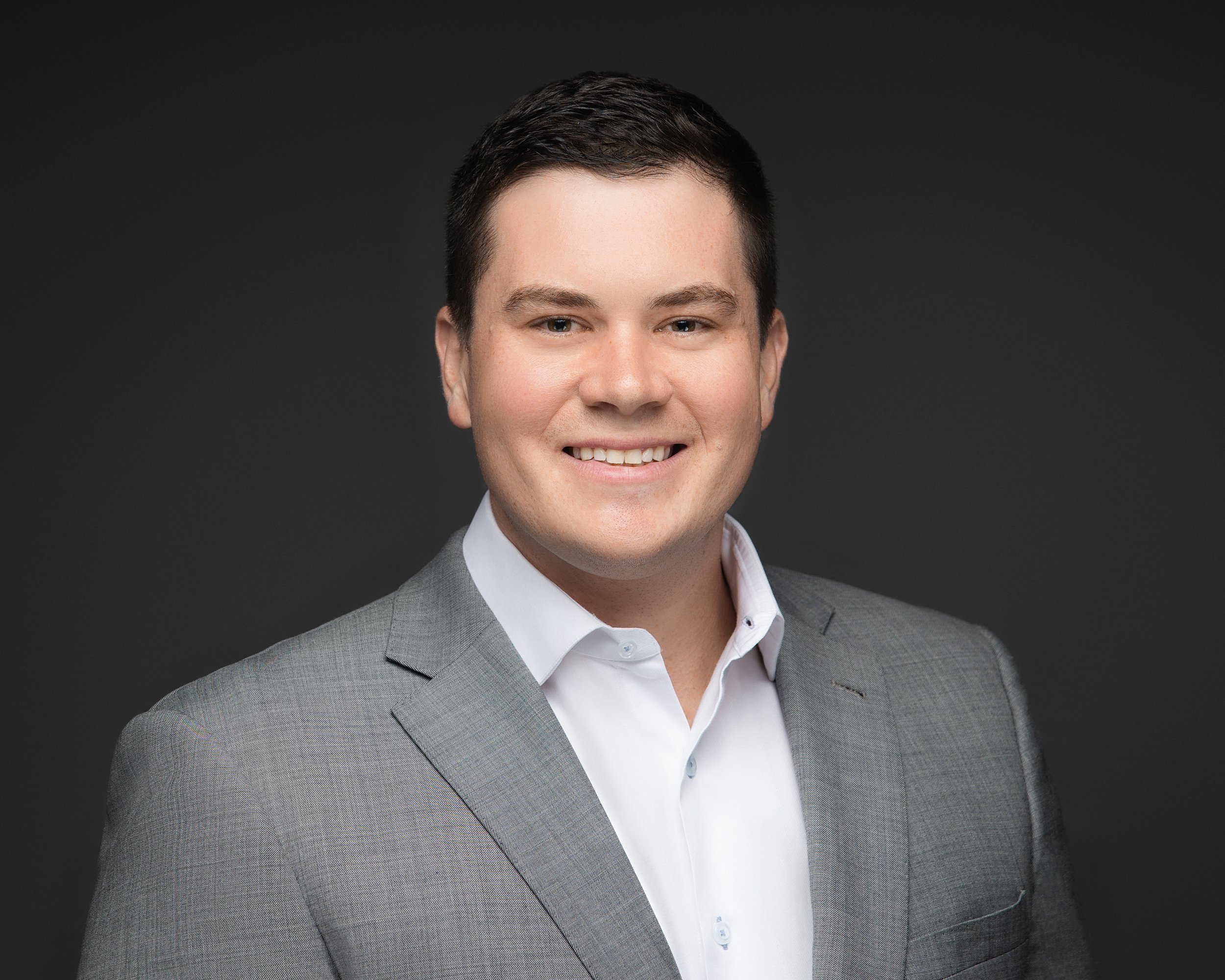 A young man with short dark hair, wearing a light gray suit and white shirt, smiling, in front of a dark gray background.