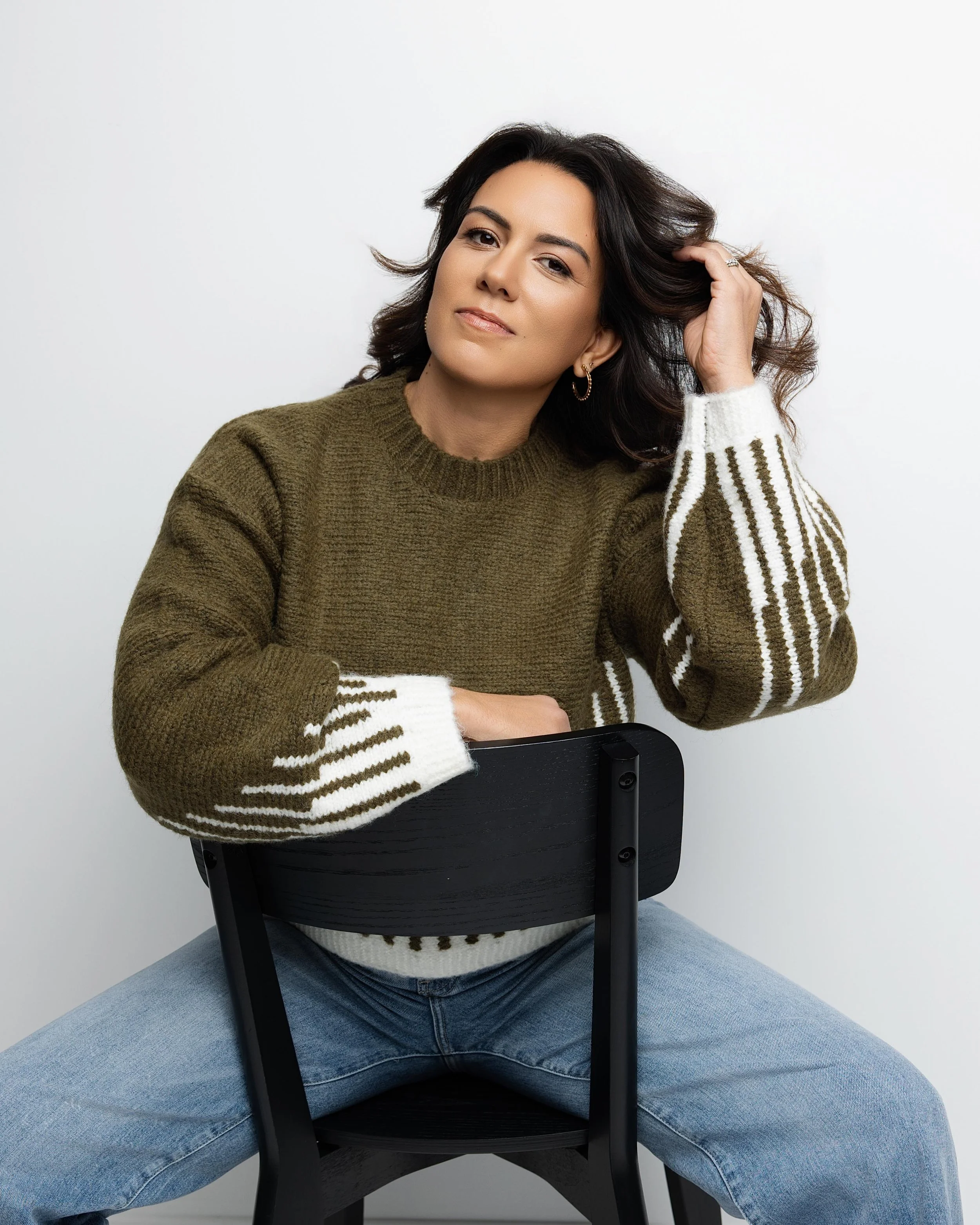 A woman with long dark hair sitting on a black chair, wearing a brown sweater with white and brown striped sleeves, and light blue jeans. She is posing with one hand touching her hair and the other resting on the back of the chair against a plain whi