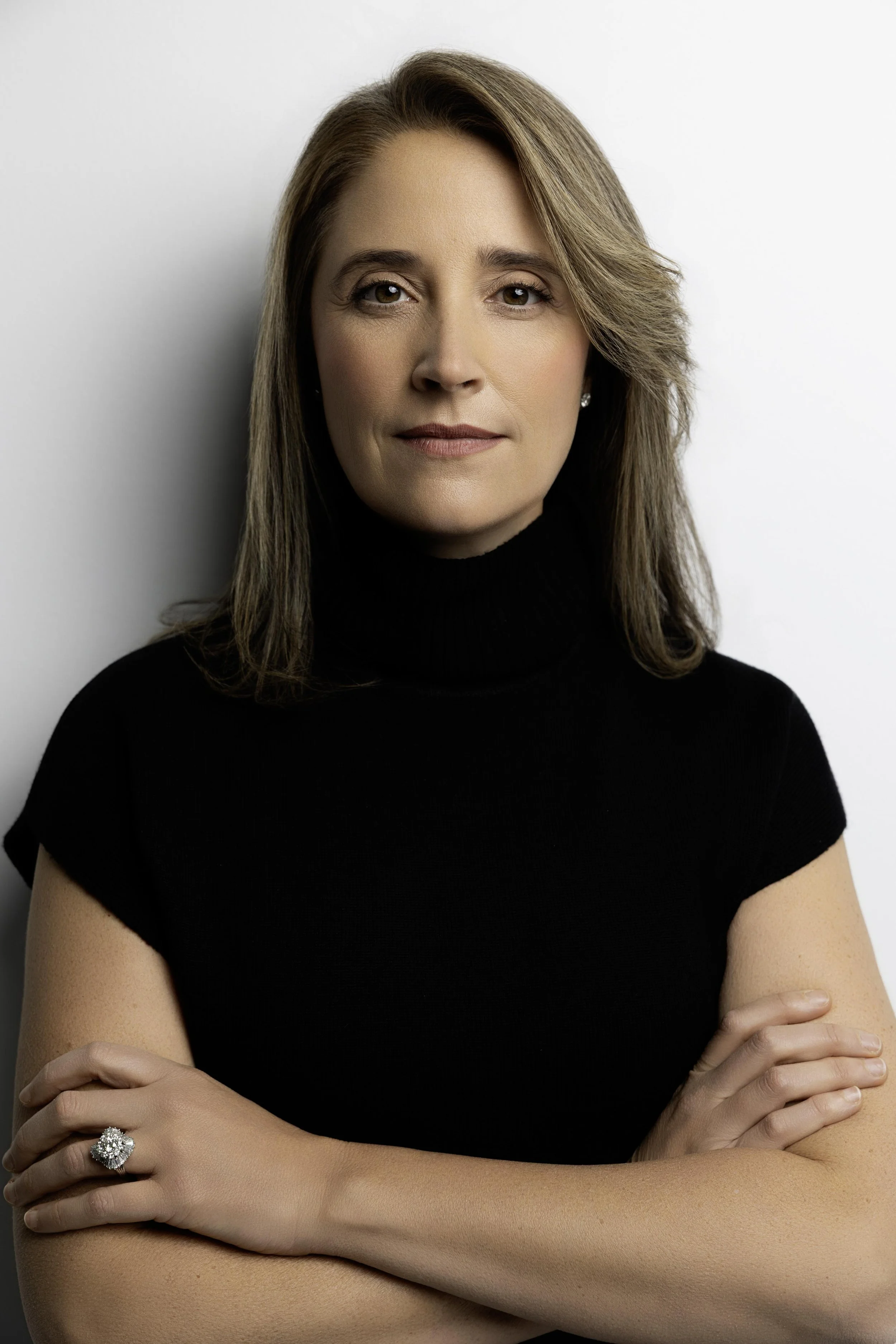 A woman with shoulder-length light brown hair wearing a black turtleneck sweater, crossed arms, standing against a plain white background.