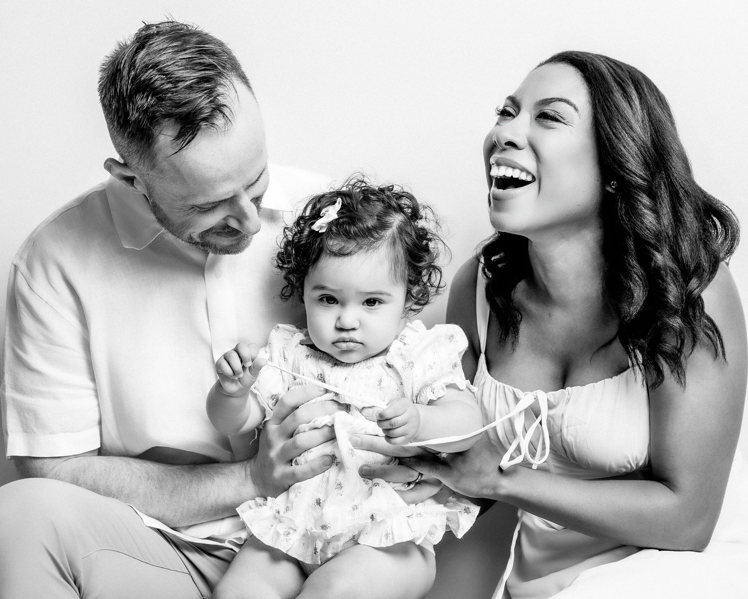 A black and white photo of a smiling woman with dark wavy hair holding a young girl with curly hair and a bow, with a man with short hair and a beard looking at the girl, all appearing to be happy.