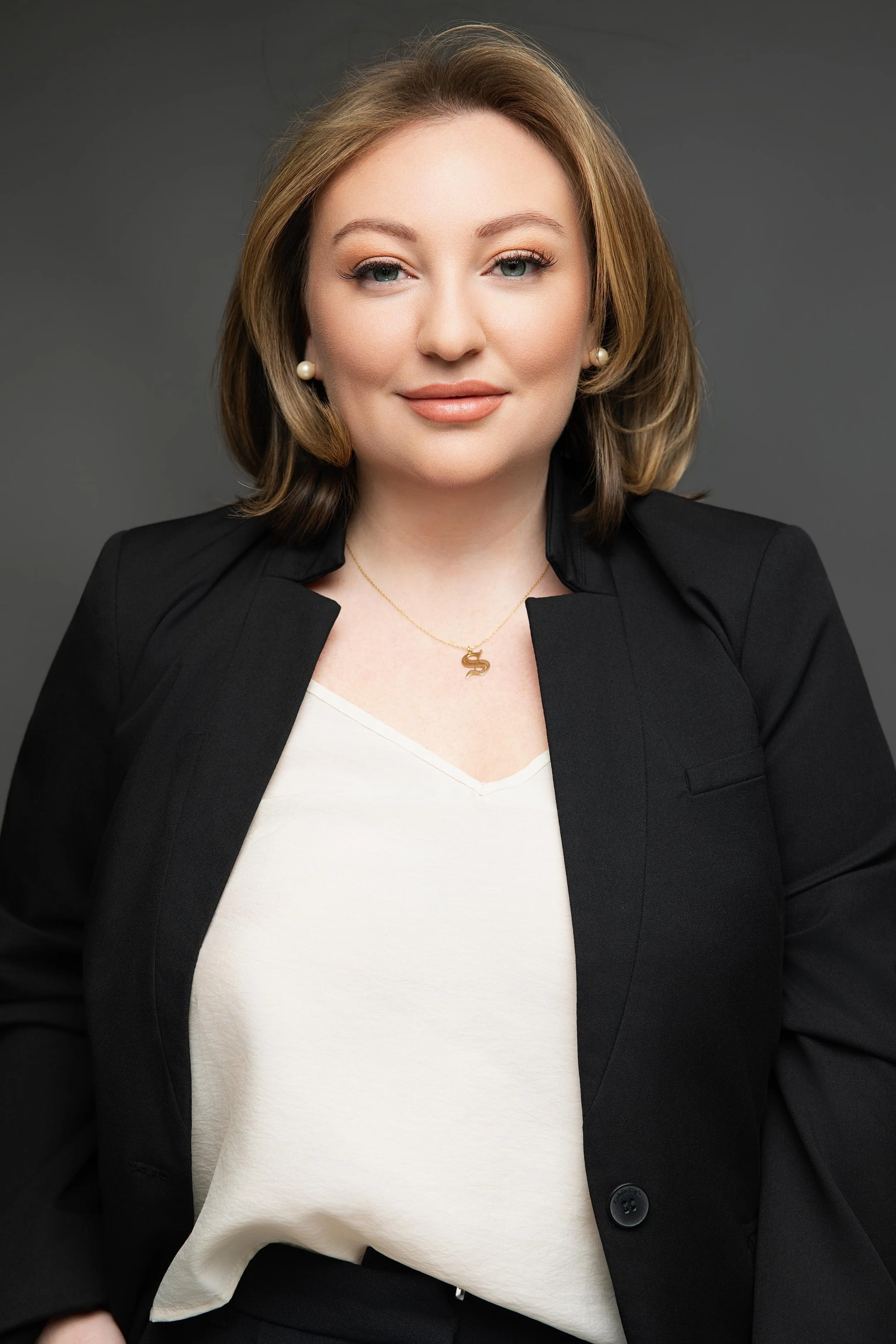 A woman with long, wavy blonde hair smiling, seated against a dark background, wearing a white blazer, a dark top, and jewelry.