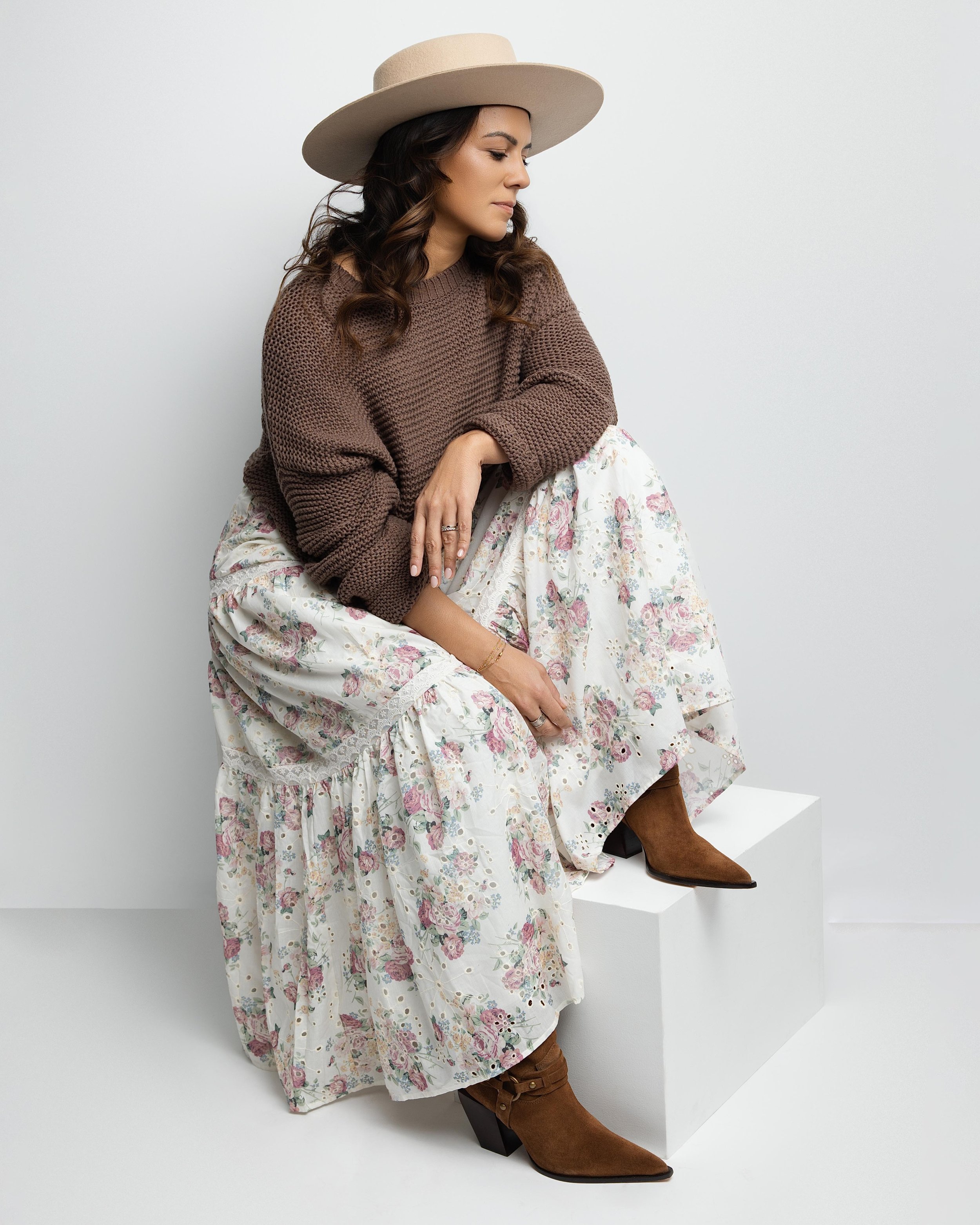 A woman wearing a beige wide-brim hat, a brown knitted sweater, a floral white skirt, and brown ankle boots, sitting on a white platform against a plain white background.