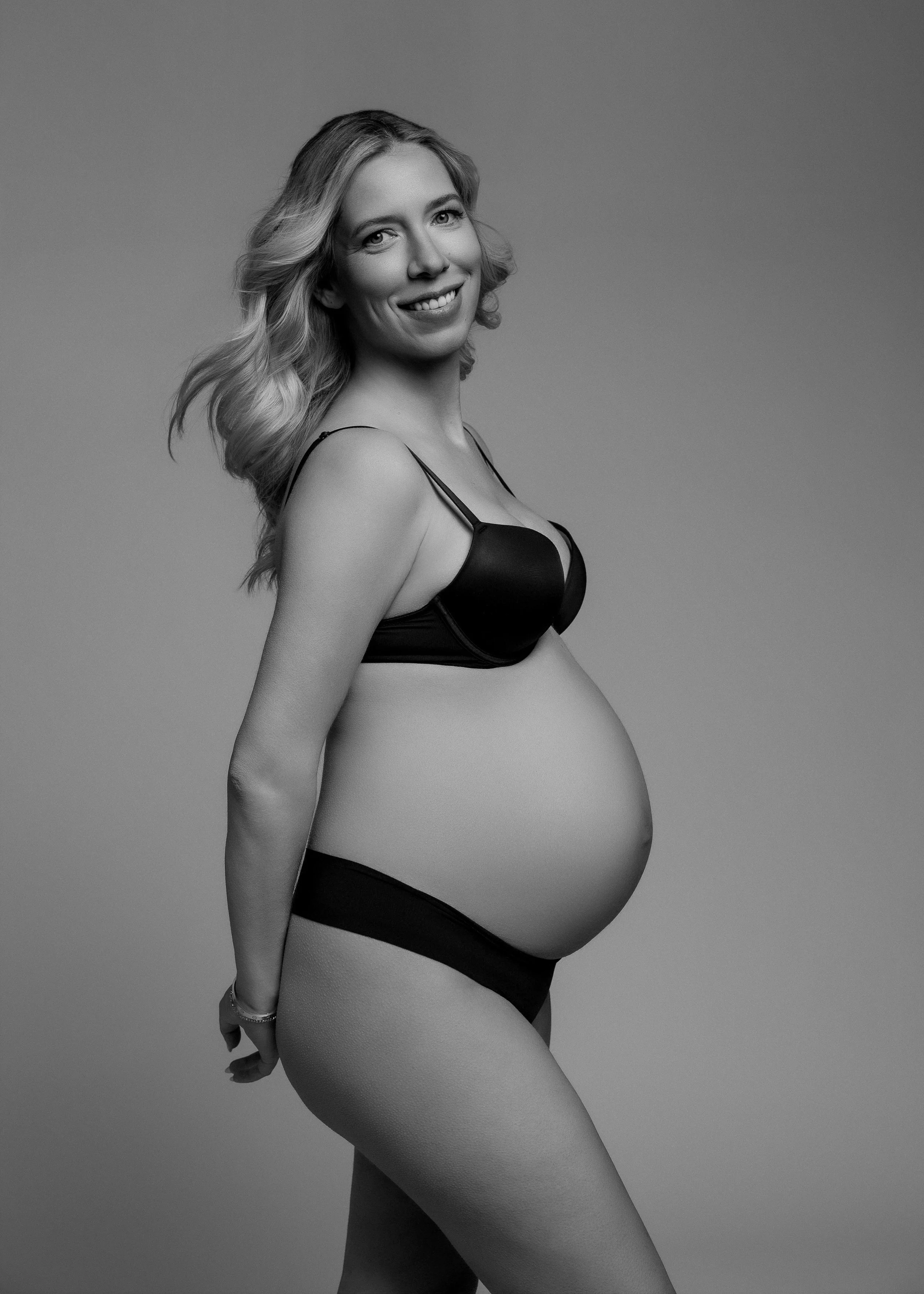 Black-and-white photo of a pregnant woman with wavy hair, smiling, wearing black lingerie, standing against a plain background.