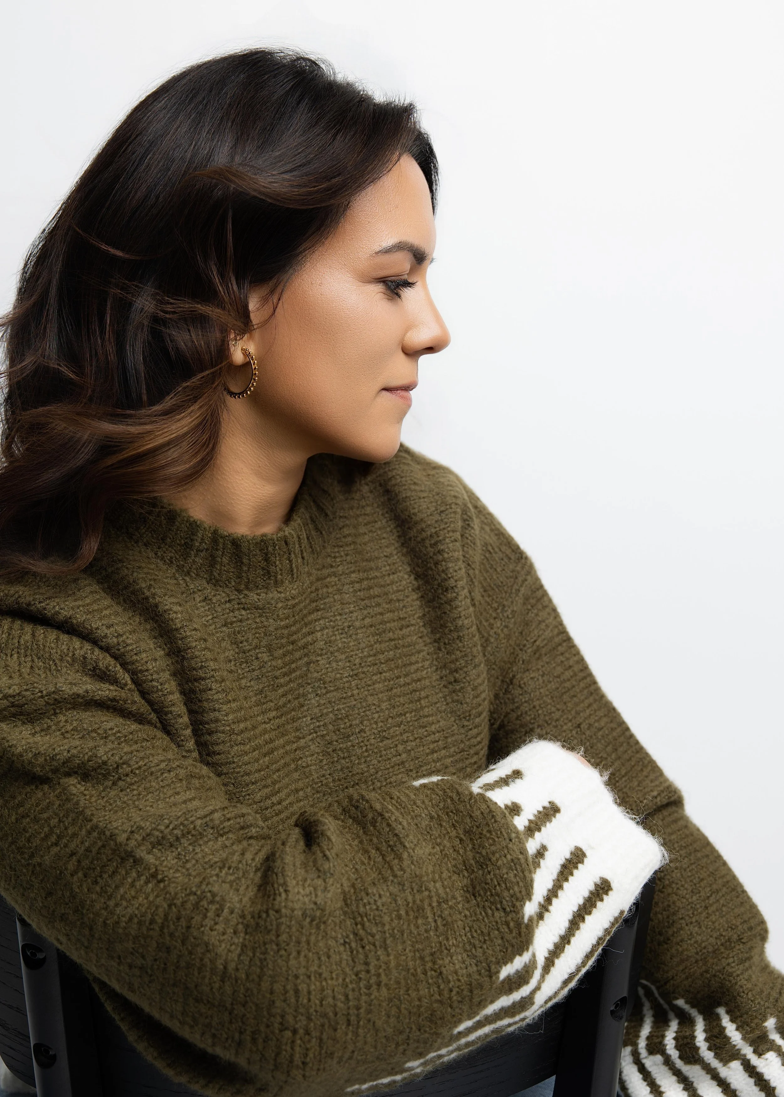 Side profile of a young woman with brown hair wearing a brown knitted sweater with white and brown striped sleeves, sitting on a black chair against a white background.