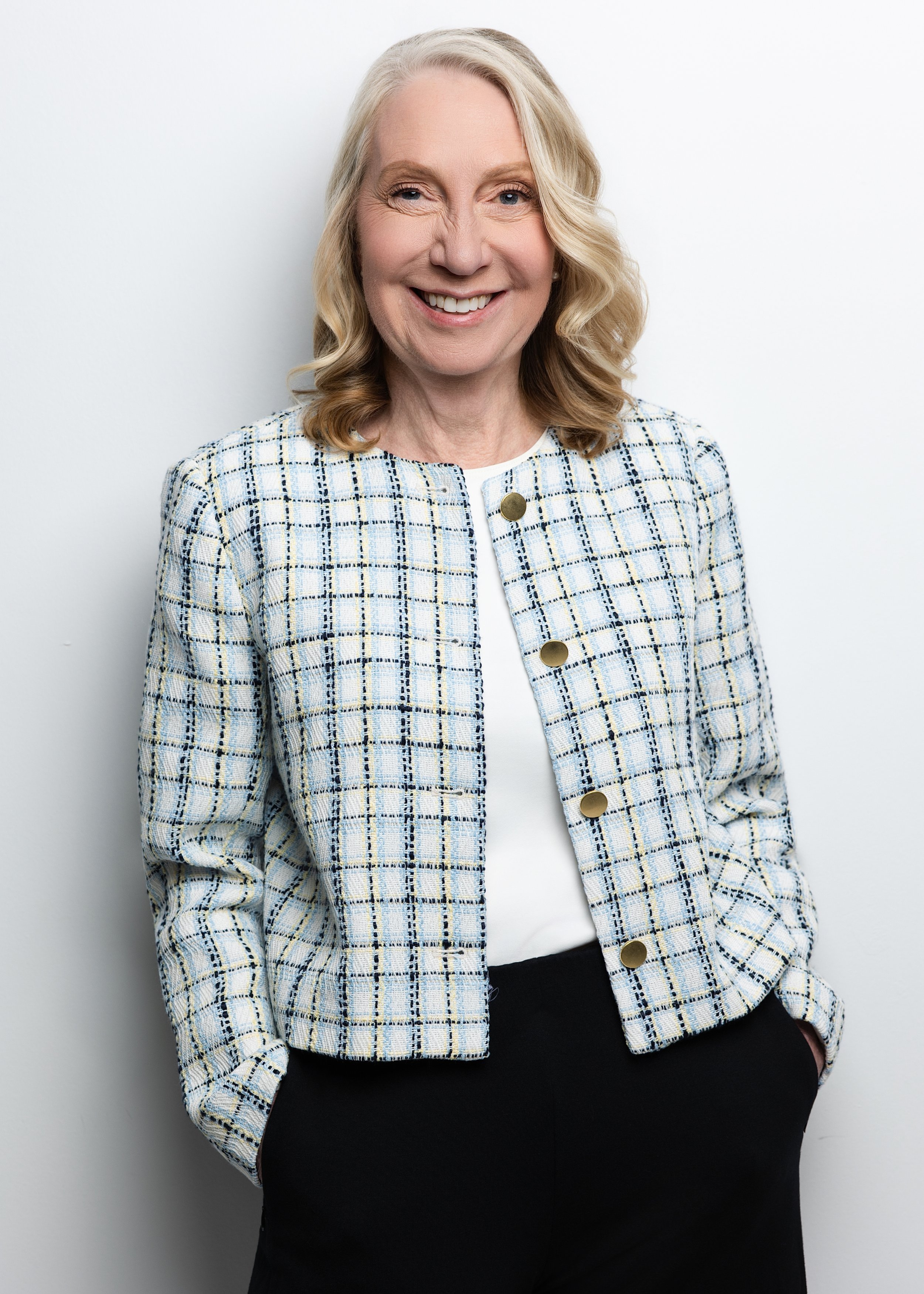 A middle-aged woman with blonde hair, wearing a light-colored plaid blazer with gold buttons over a white top, smiling confidently with one hand in her pocket against a plain white background.