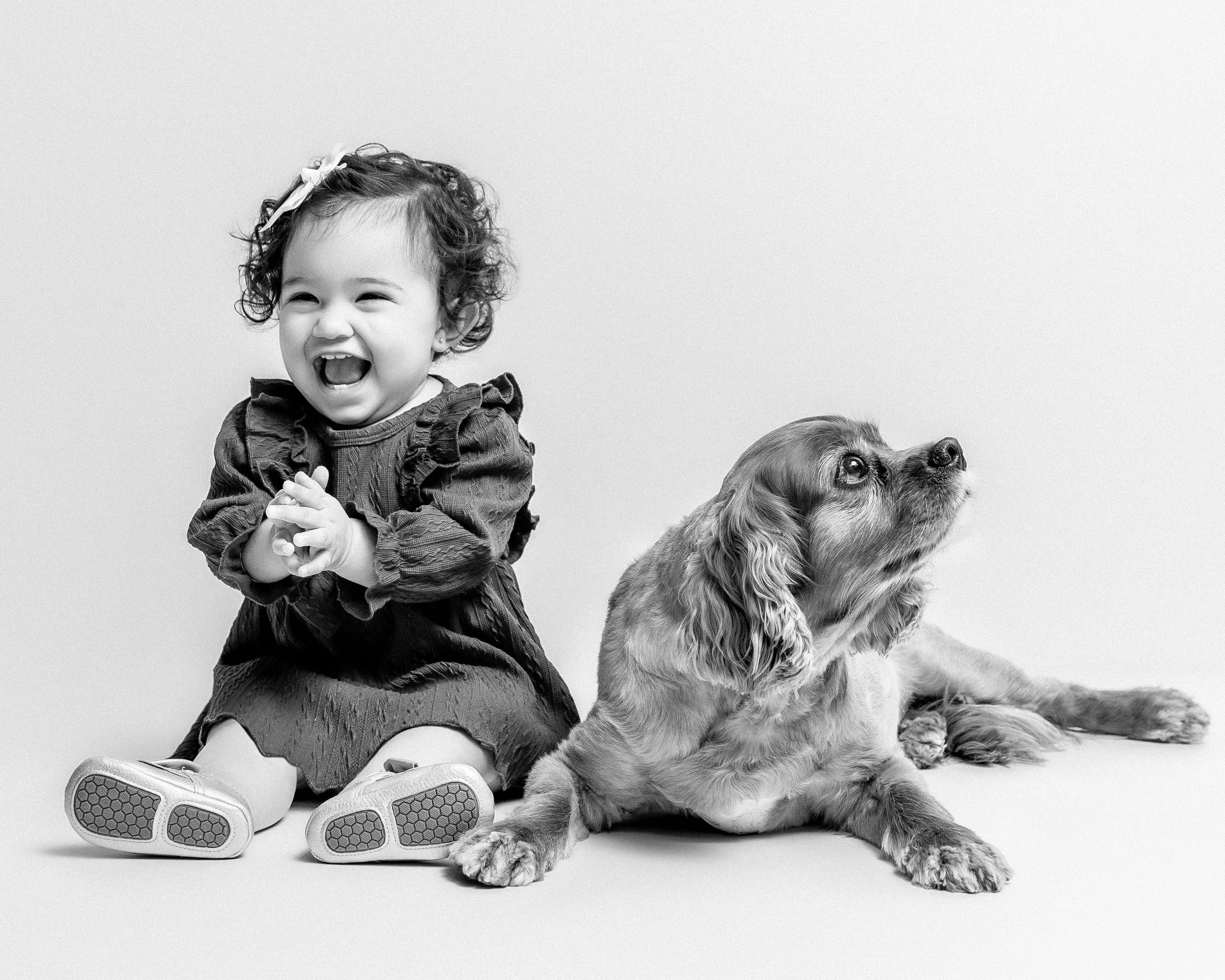 A young girl with curly hair, wearing a ruffled dress and sitting on the floor, smiling with her eyes closed. A dog, lying down next to her, looking upward.