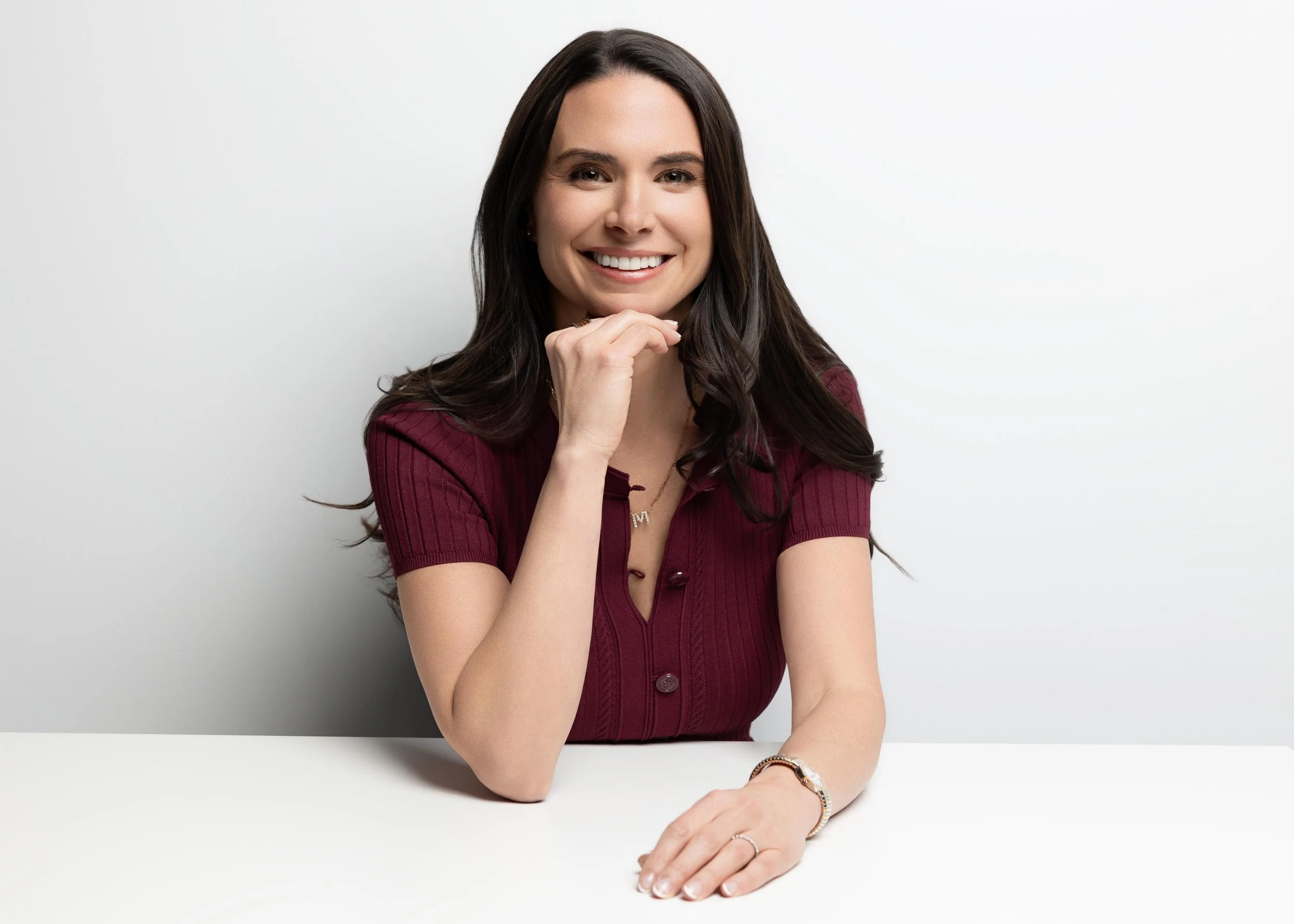 A woman with long dark hair wearing a maroon short-sleeved button-up top, sitting at a white table, smiling with her chin resting on her right hand, against a plain white background.