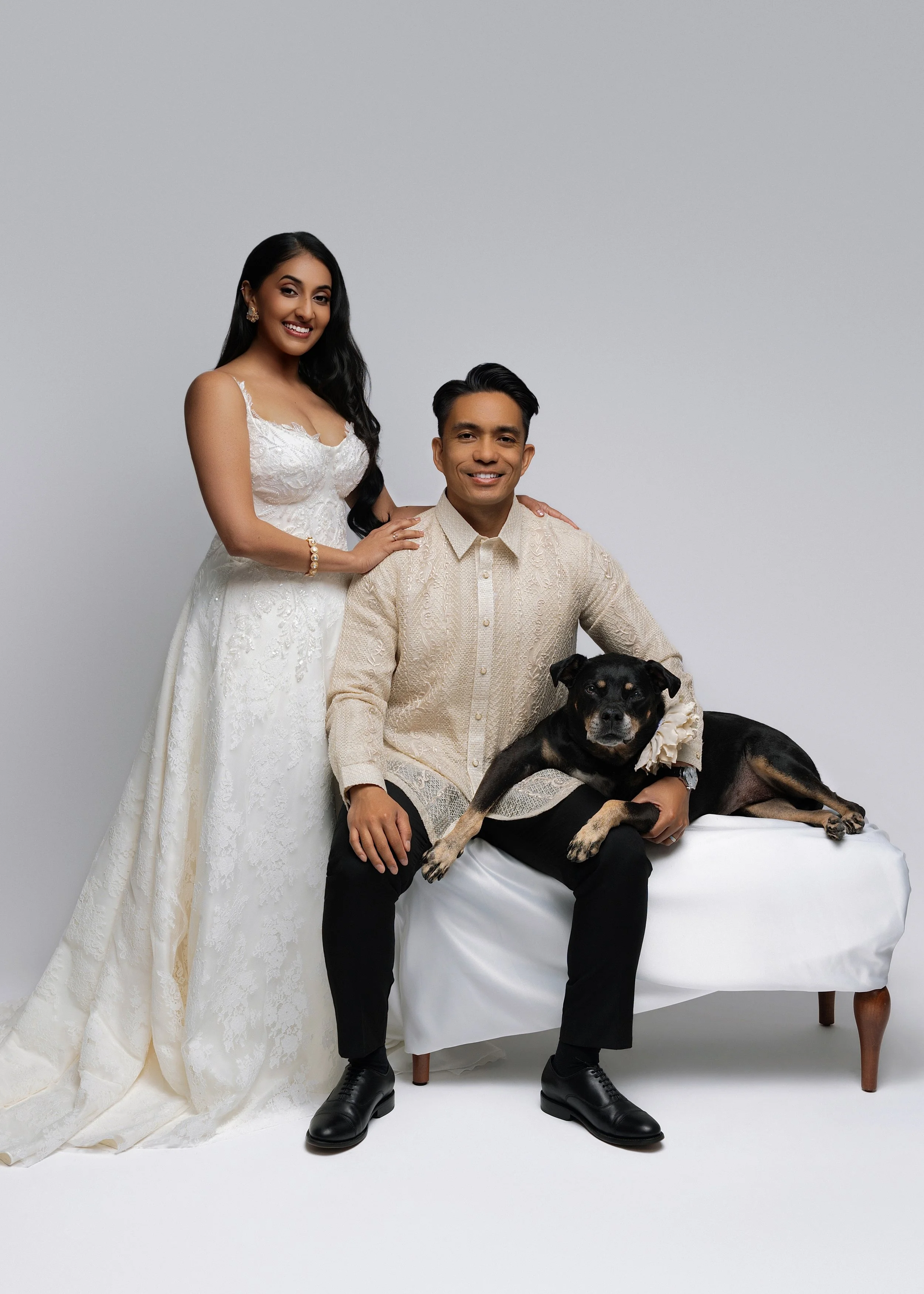 A couple dressed in wedding attire, with the woman in a white wedding dress and the man in a beige traditional outfit, sitting on a small white table. The woman has long black hair and is standing behind the seated man, with her hand on his shoulder.