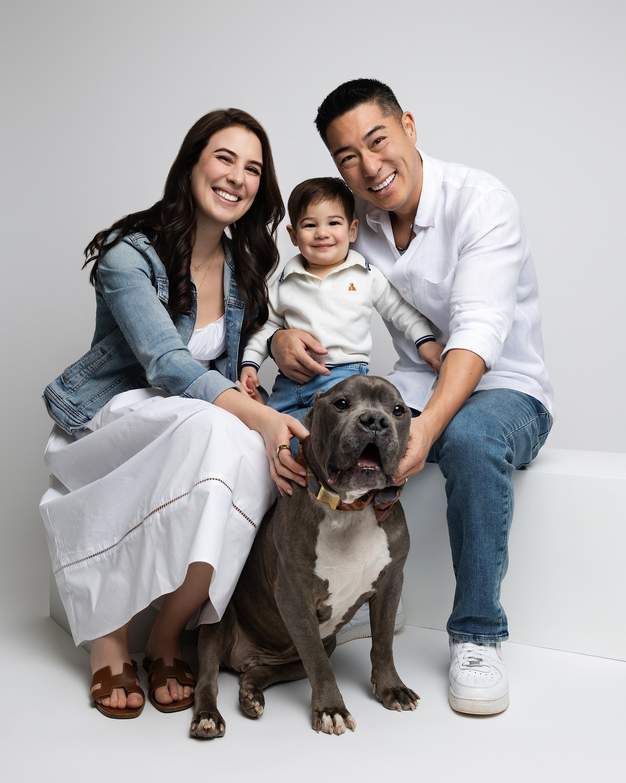 A happy multiracial family with a dog, smiling and sitting together against a plain white background.