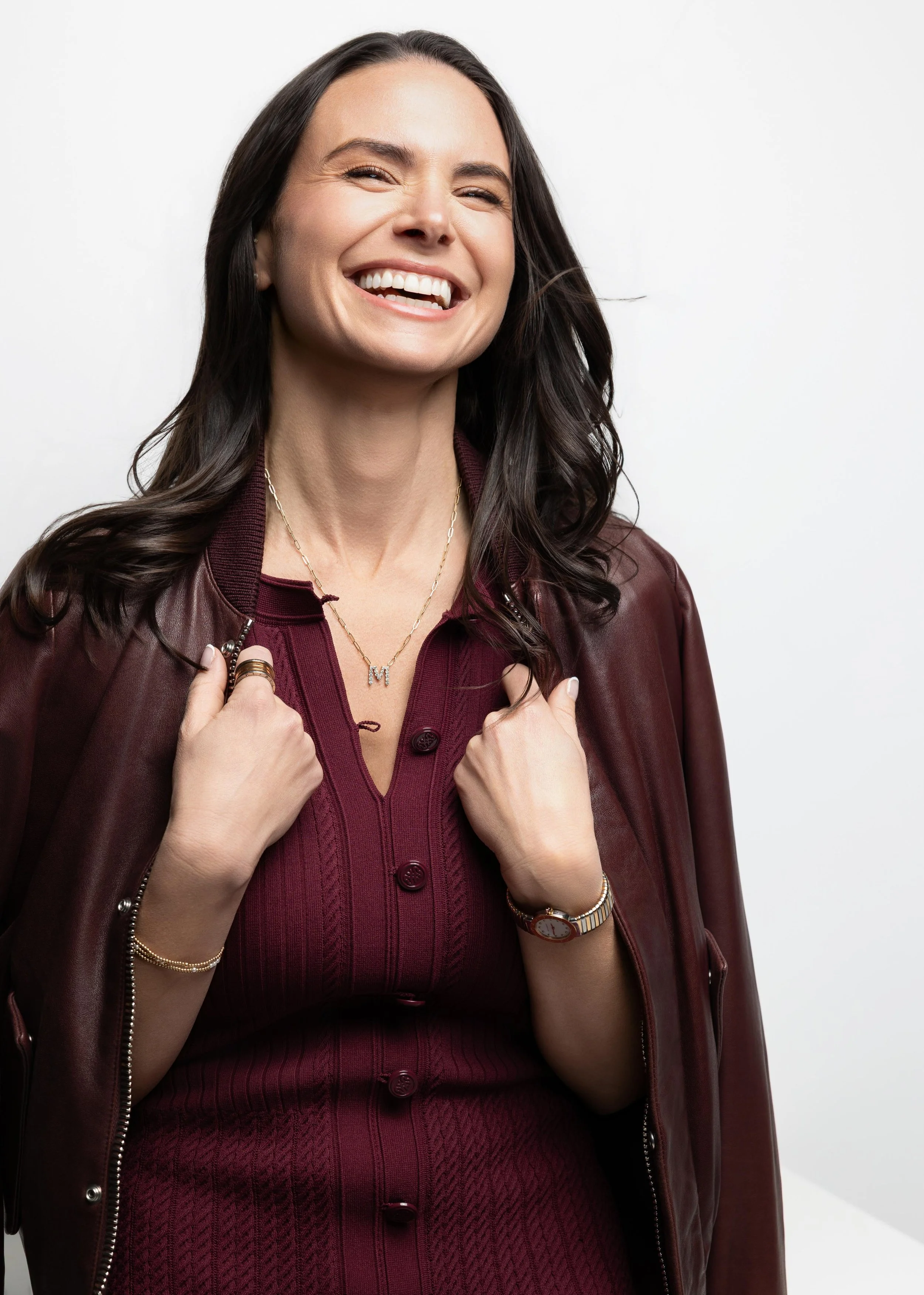 A woman with long dark hair smiling and showing her teeth, wearing a burgundy button-up sweater, a matching jacket draped over her shoulders, a watch, gold bracelets, and a necklace with a letter 'M' pendant.
