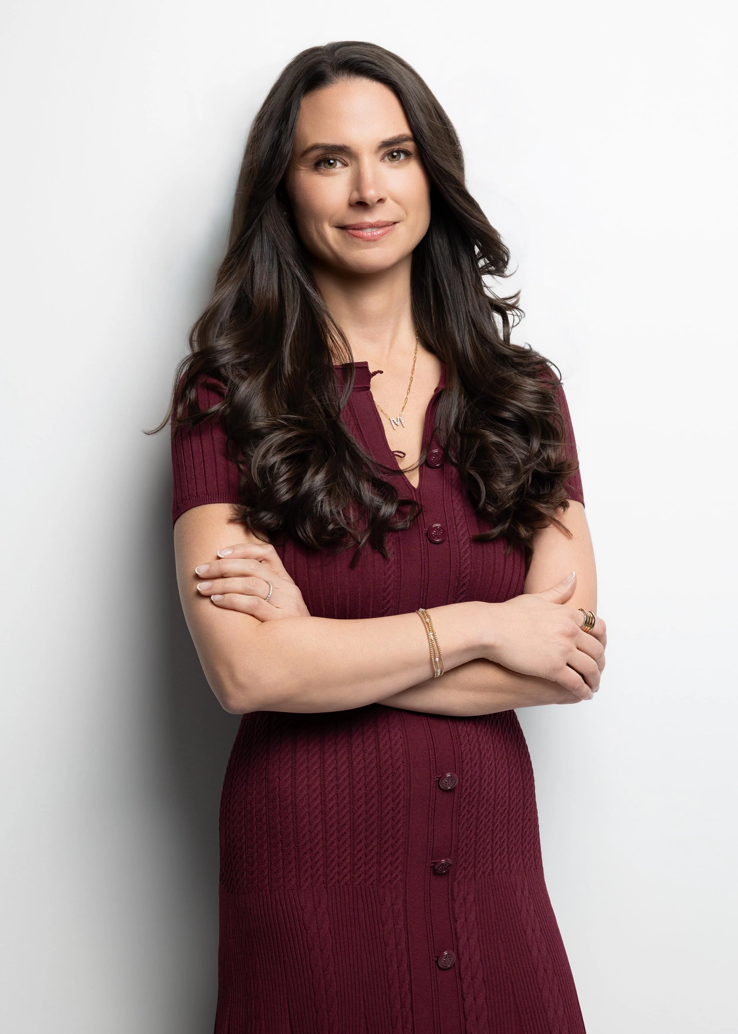 A woman with long, dark, wavy hair wearing a burgundy knitted dress with buttons, silver jewelry, and crossed arms against a plain white background.