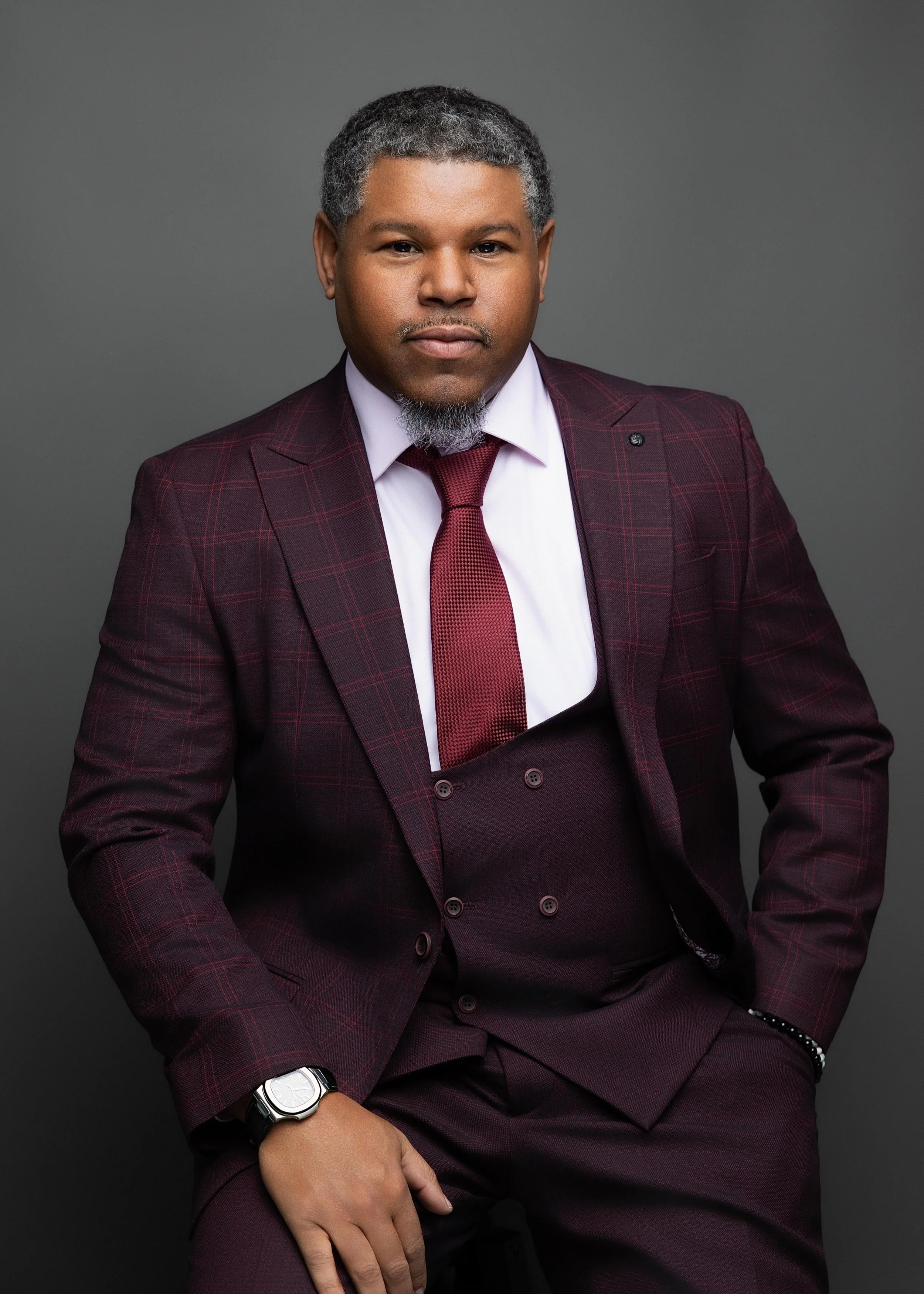 A man dressed in a maroon plaid suit with a white shirt, burgundy tie, and matching pants, standing against a gray background.