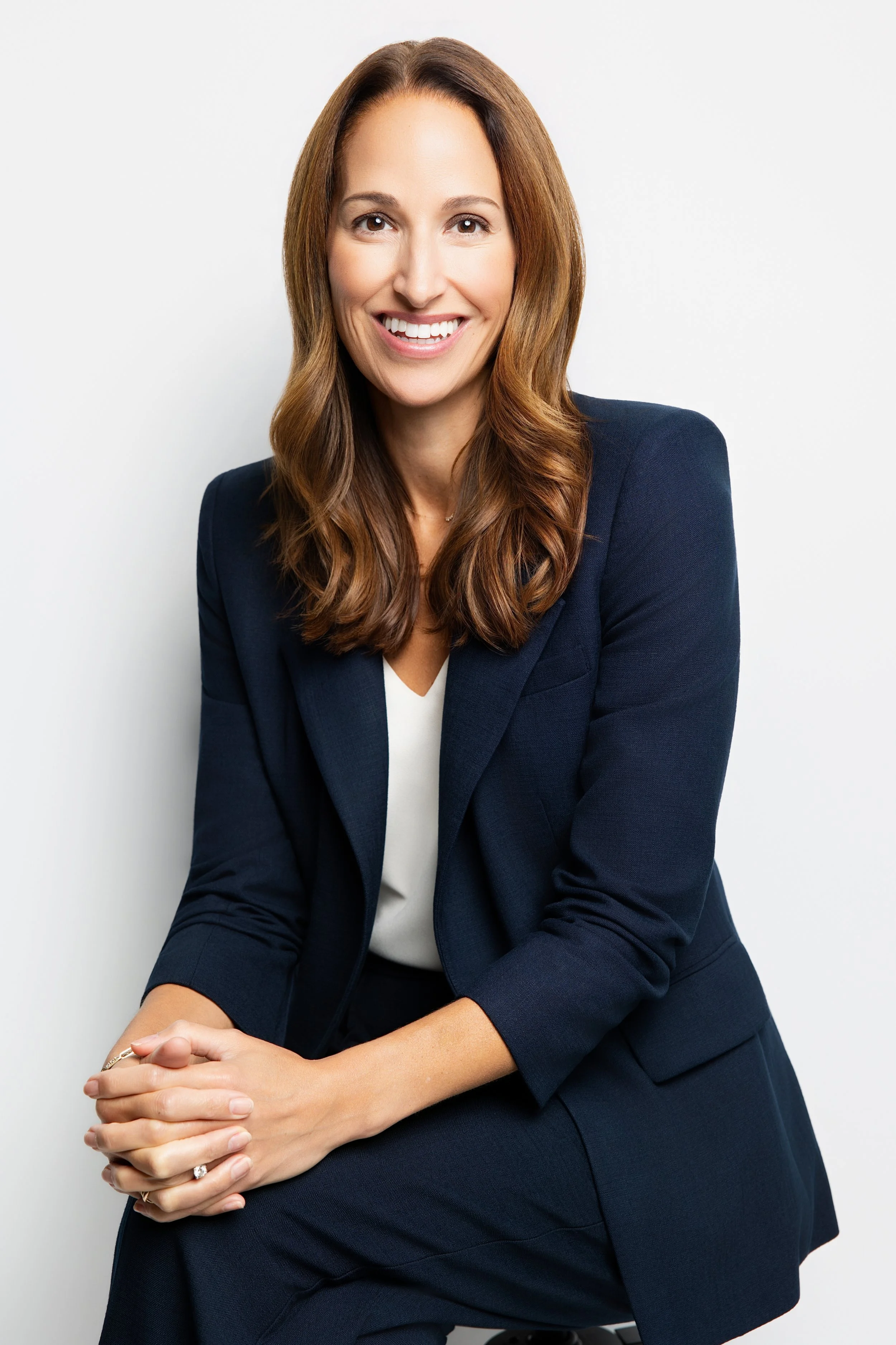 Portrait of a woman with long brown hair smiling, wearing a navy blazer and white top, sitting against a plain white background.