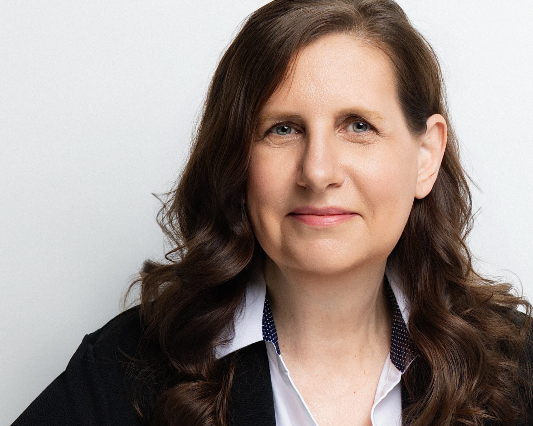 Close-up portrait of a woman with dark brown, shoulder-length hair and blue eyes, wearing a white collared shirt and black blazer, against a plain light background.