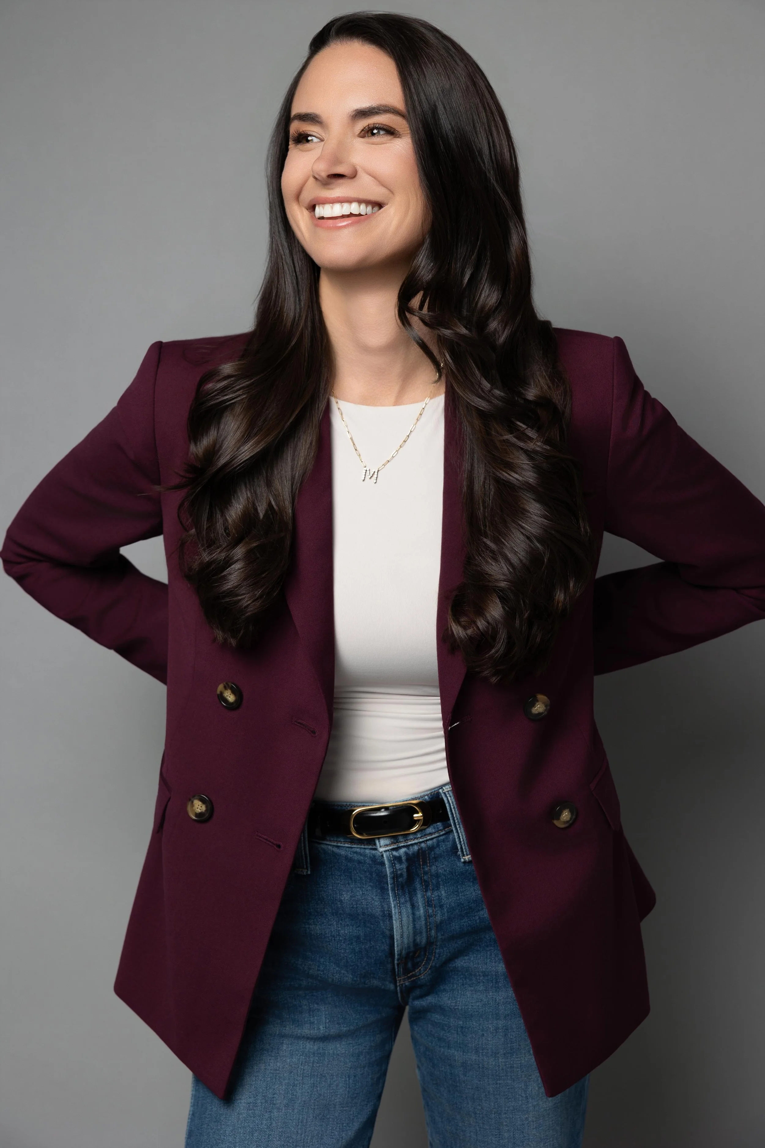 A woman with long dark wavy hair smiling, wearing a white top, a dark red blazer, and blue jeans, standing against a gray background.
