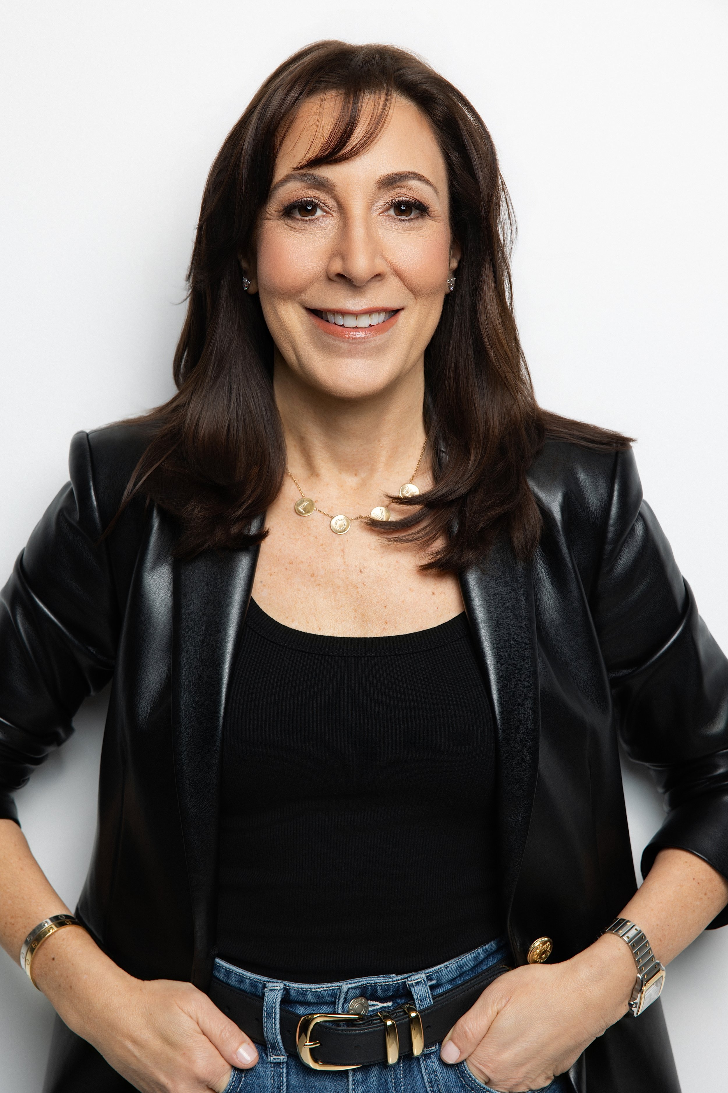 A woman with shoulder-length brown hair, wearing a black leather jacket over a black top, smiling with her hands on her hips, standing against a white background.