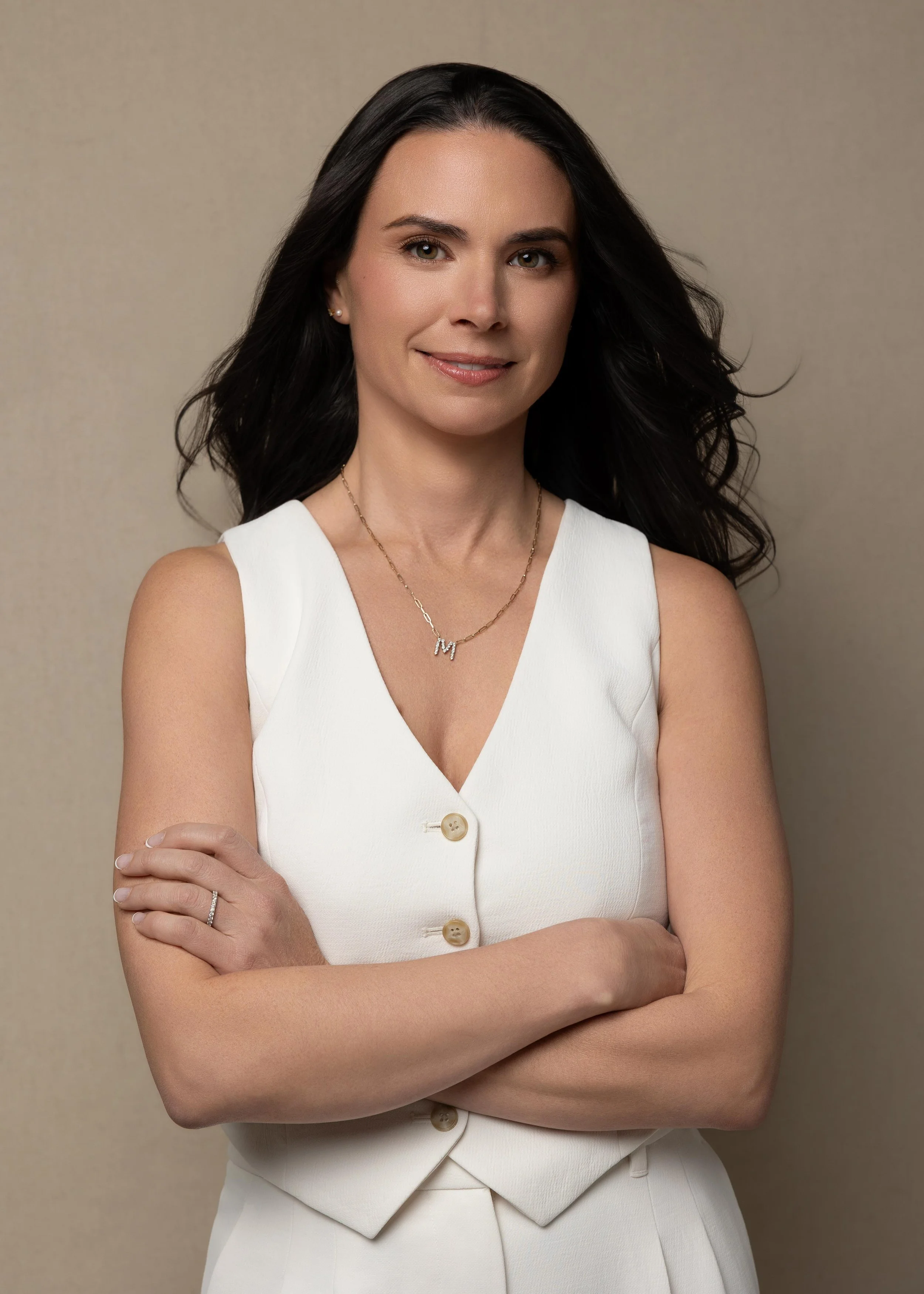 A woman with long dark hair, wearing a white sleeveless top with buttons, gold necklace, and a ring on her left hand, posing against a beige background.