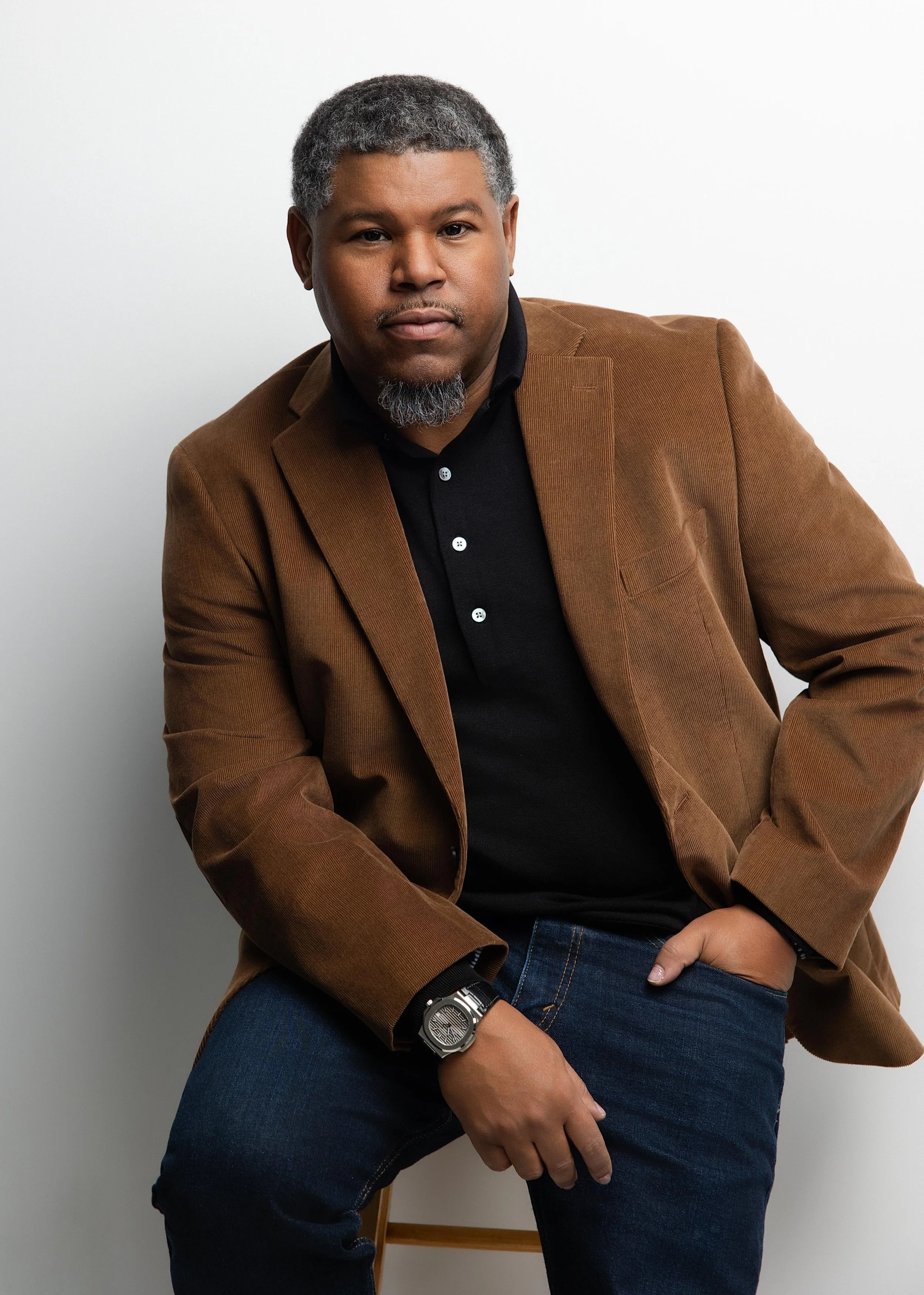 A man with short gray hair and a salt-and-pepper beard dressed in a brown blazer, black shirt, and dark jeans, seated in front of a plain white background.