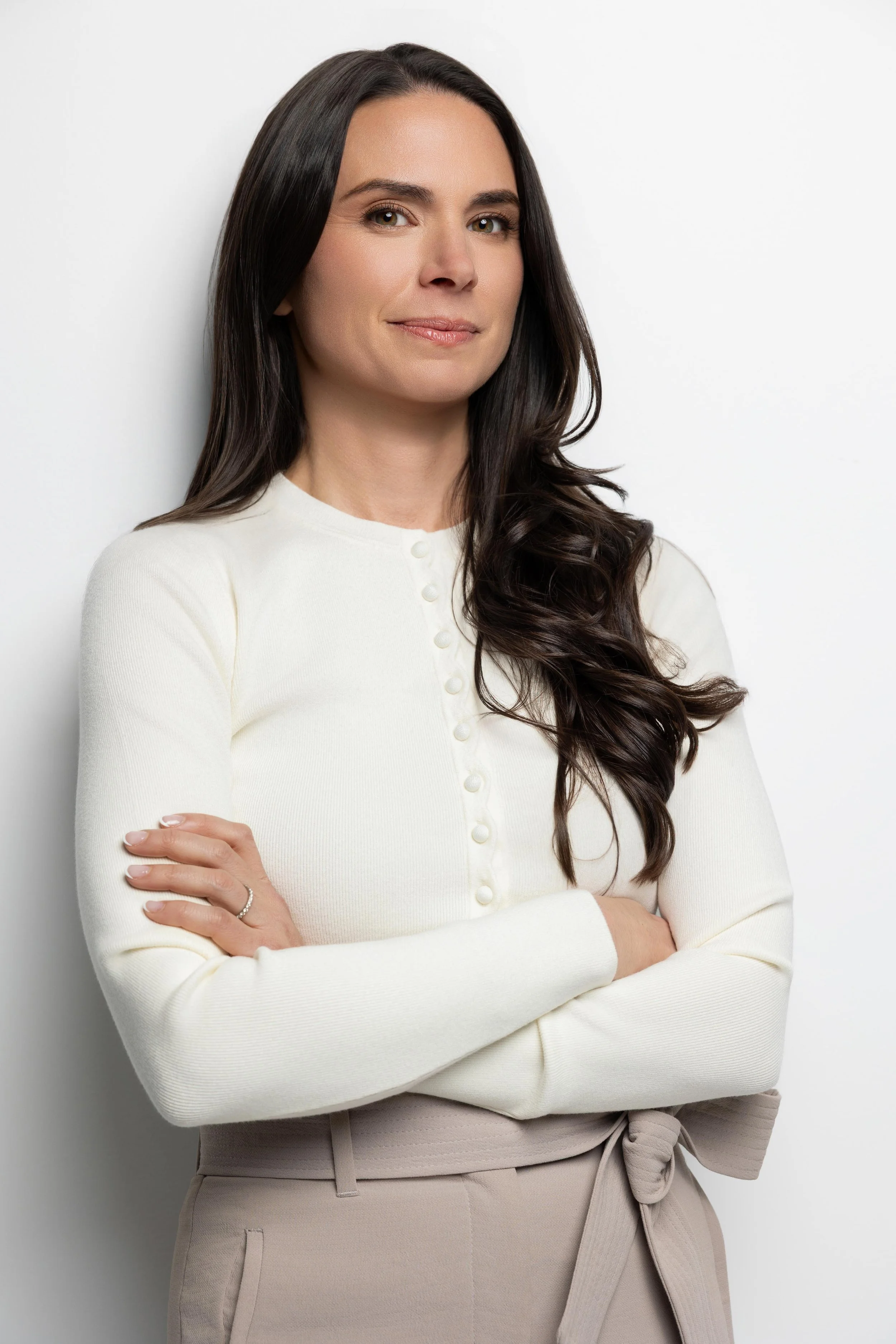 A woman with long dark hair, dressed in a cream-colored top with buttons and light beige pants, standing with her arms crossed against a white background.