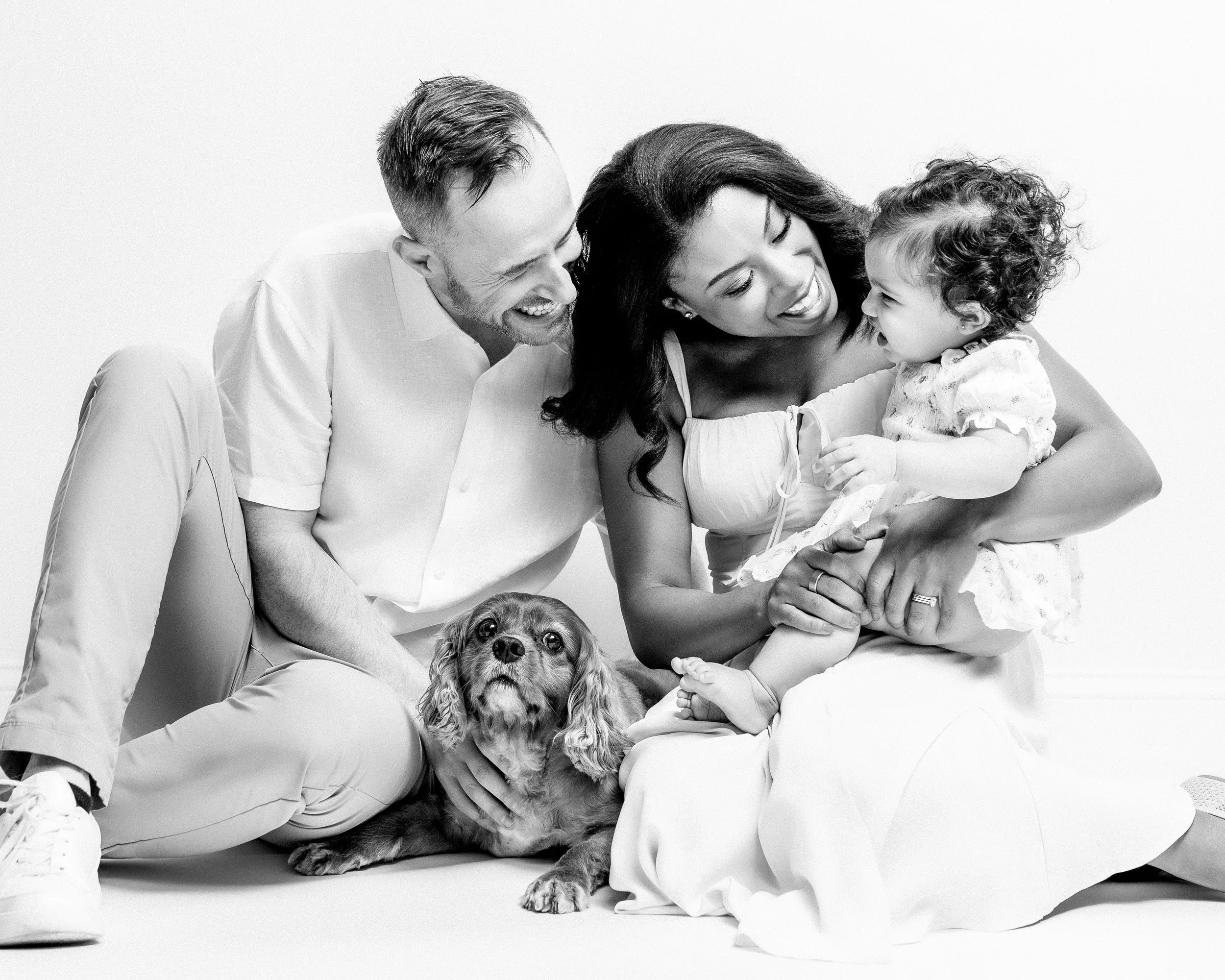A happy family of three, a man, woman, and young girl, smiles and laughs together while sitting on the floor with a dog, all in black and white.