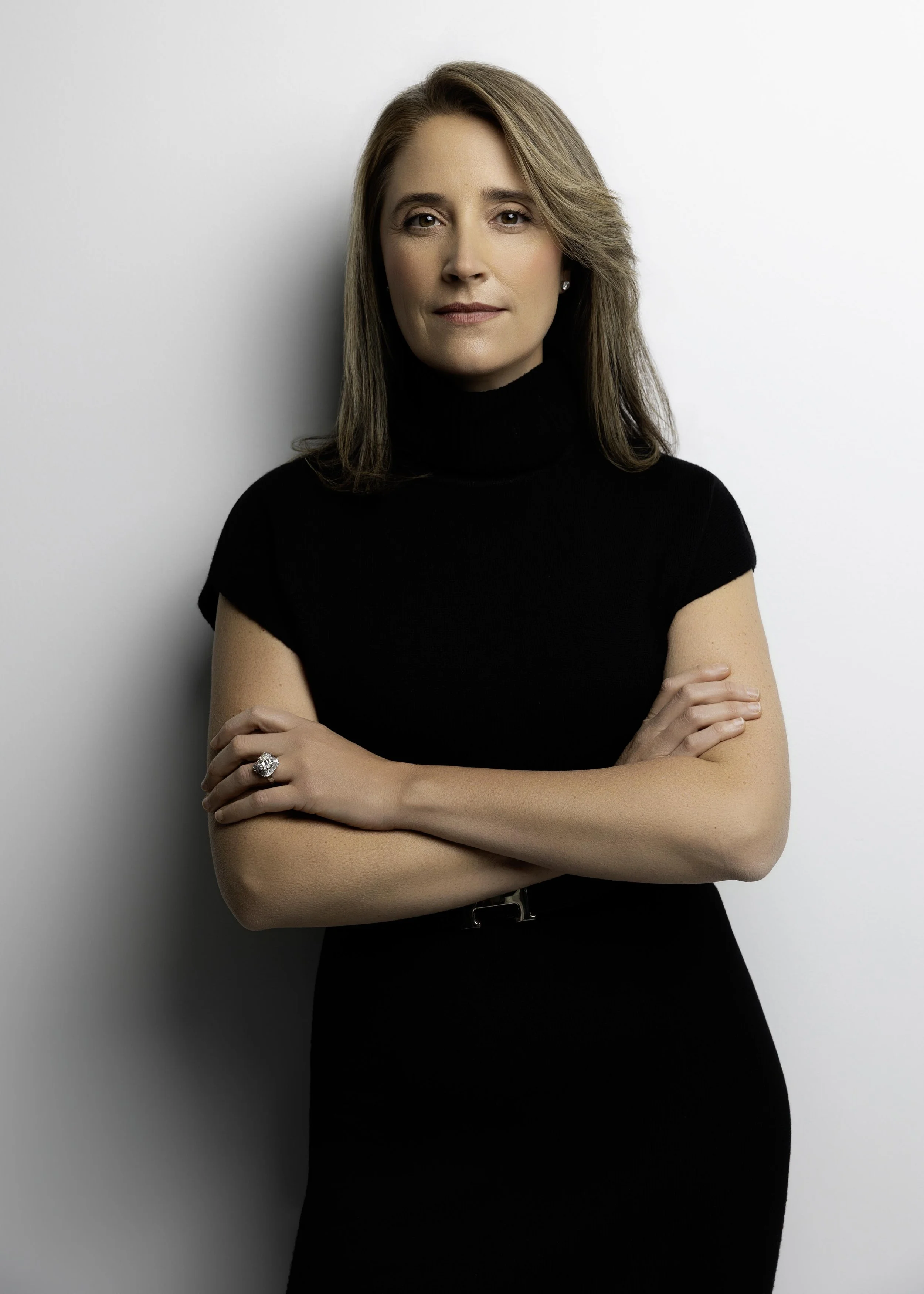 A woman with shoulder-length brown hair wearing a black turtleneck and a black dress, standing with arms crossed against a plain white background.