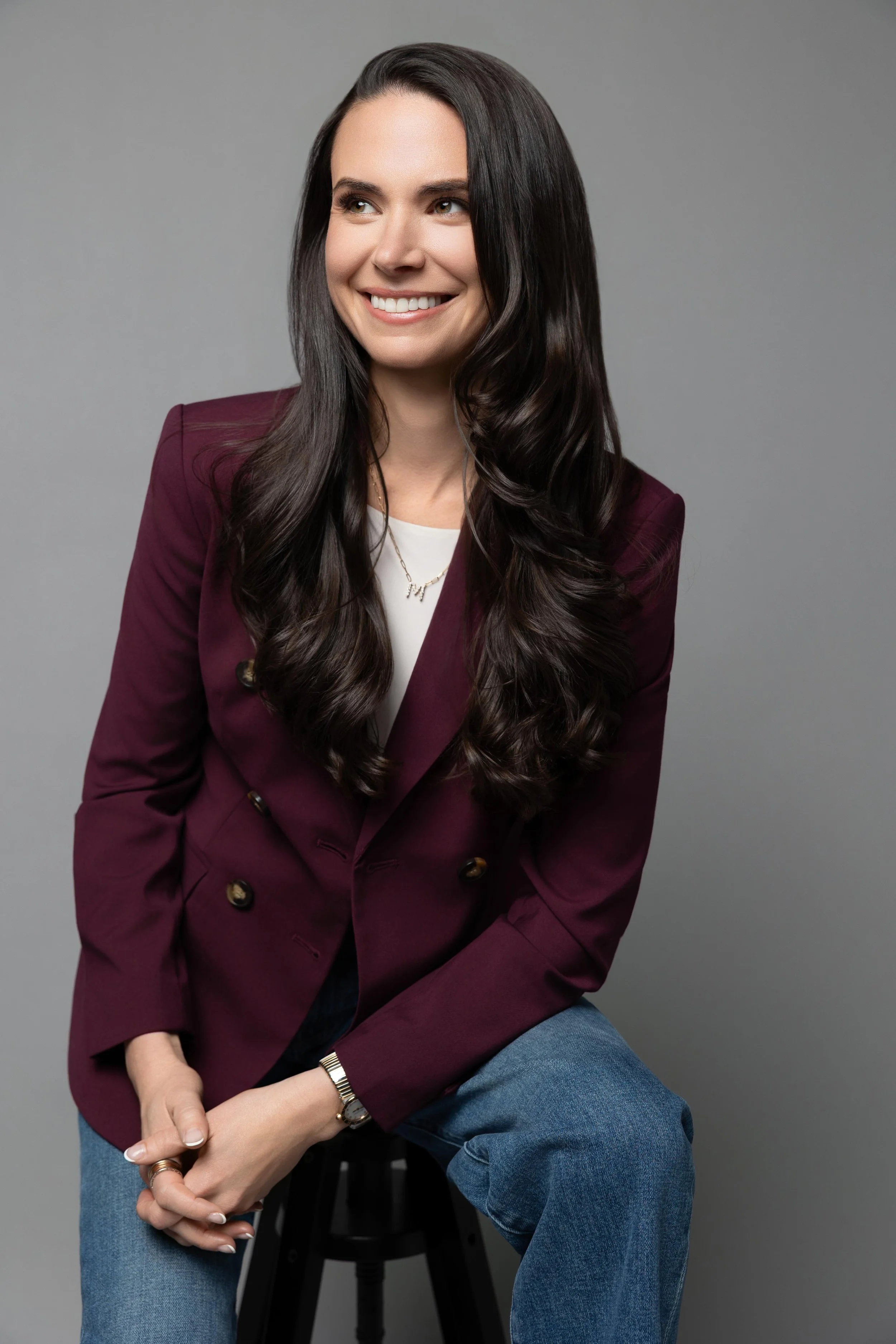 A woman with long dark hair, wearing a maroon blazer, white shirt, and blue jeans, sitting on a black stool against a gray background, smiling and looking to her left.