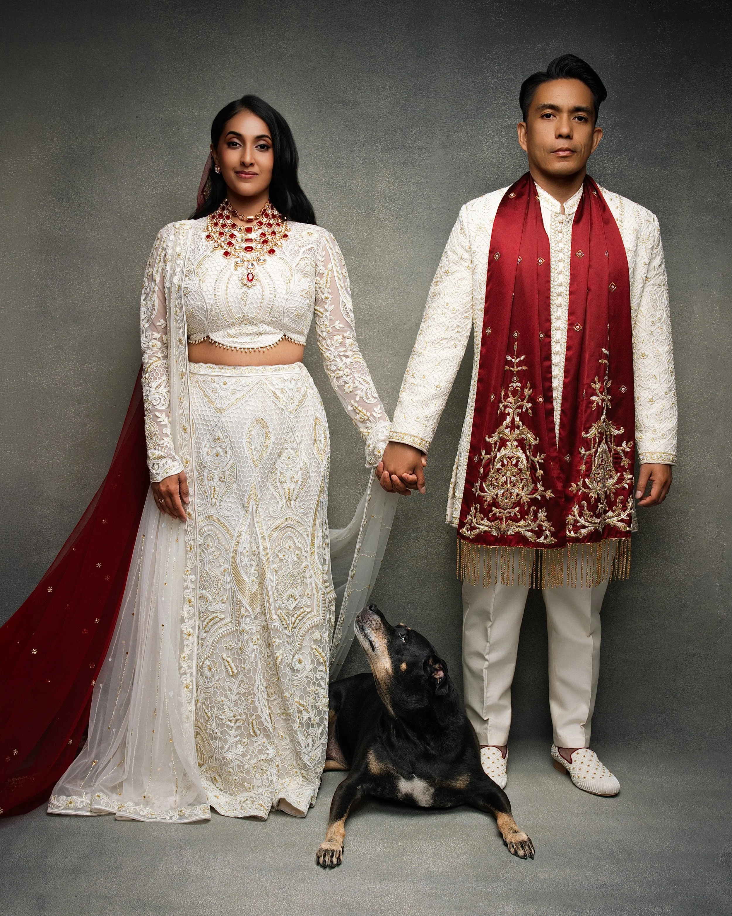 A man and woman dressed in traditional South Asian wedding attire holding hands, with a black and tan dog sitting in front. The woman is wearing a white embroidered sari with red and gold jewelry, and the man is wearing a cream embroidered sherwani w