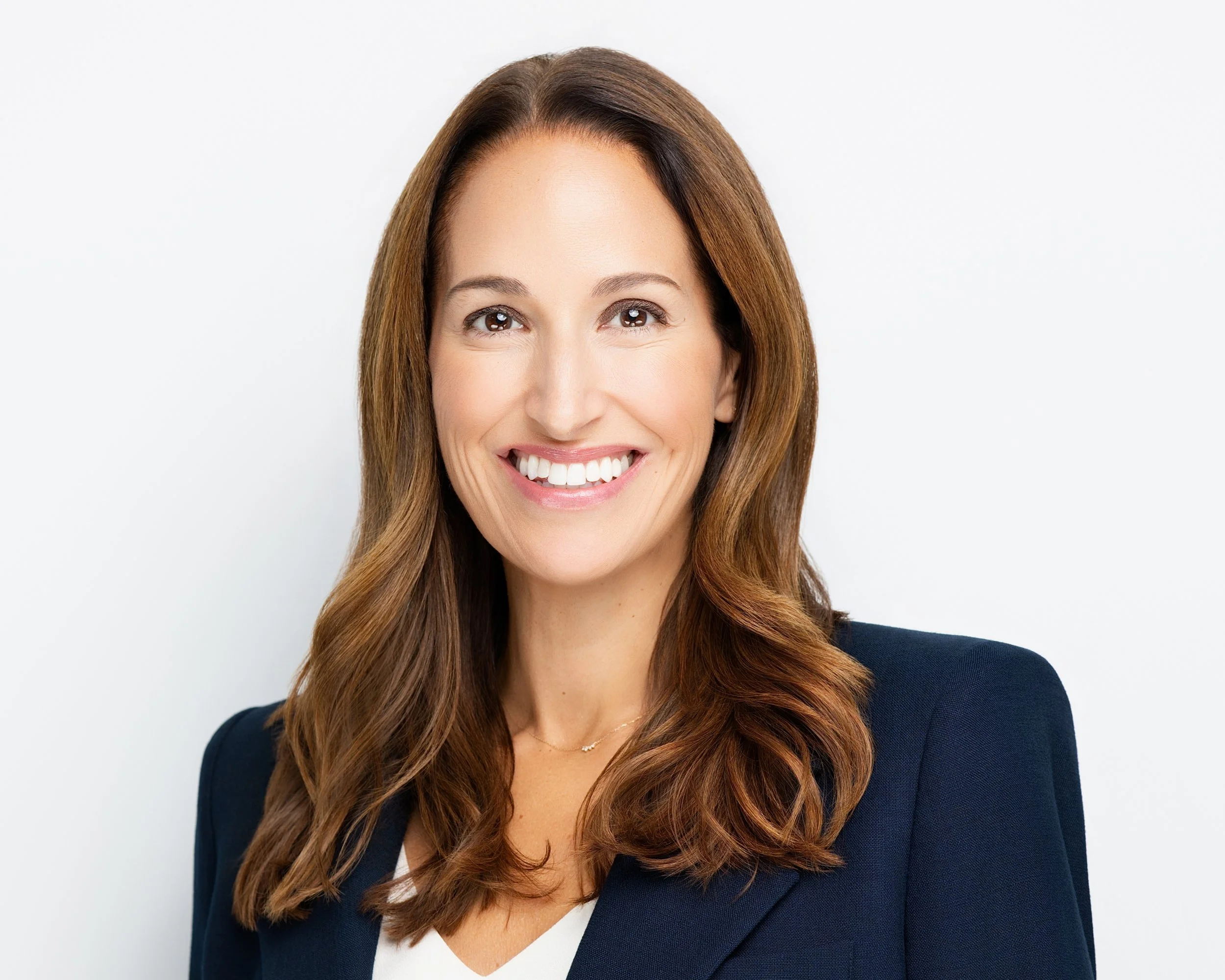 Close-up of a smiling woman with long brown hair, wearing a dark blazer and a white top, against a plain white background.