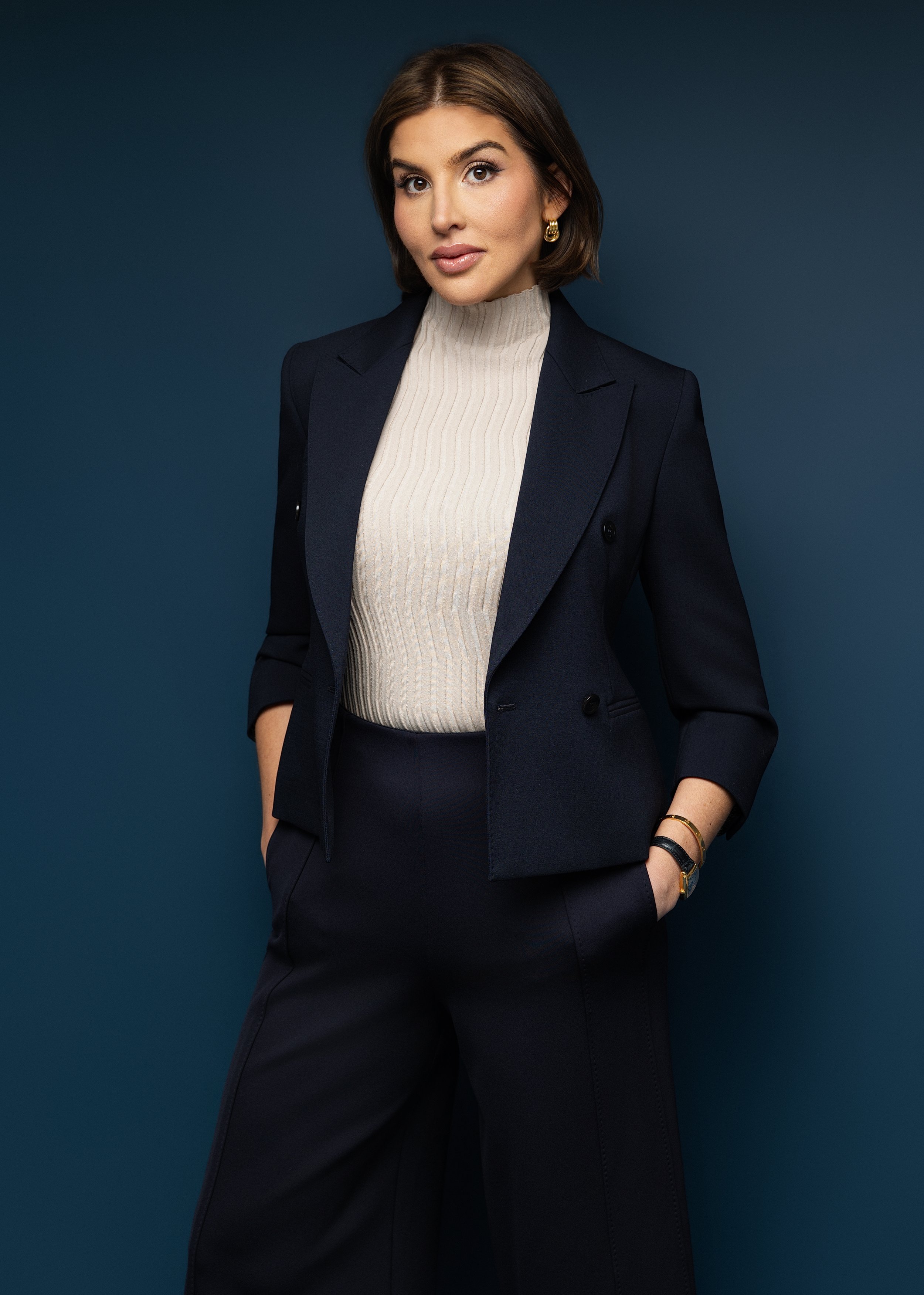 A woman in a navy pantsuit with a cream turtleneck, standing against a dark teal background, looking at the camera.